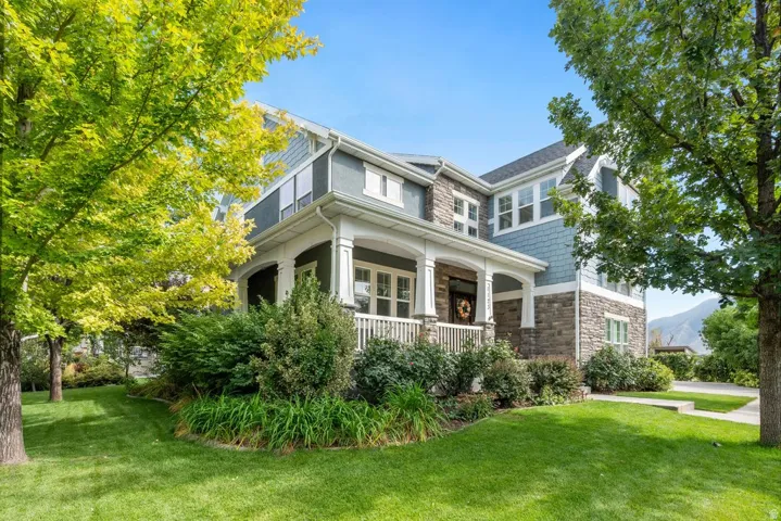 Expansive lawn with mature trees, stone and siding exterior, covered front porch with white railings, and a paved driveway