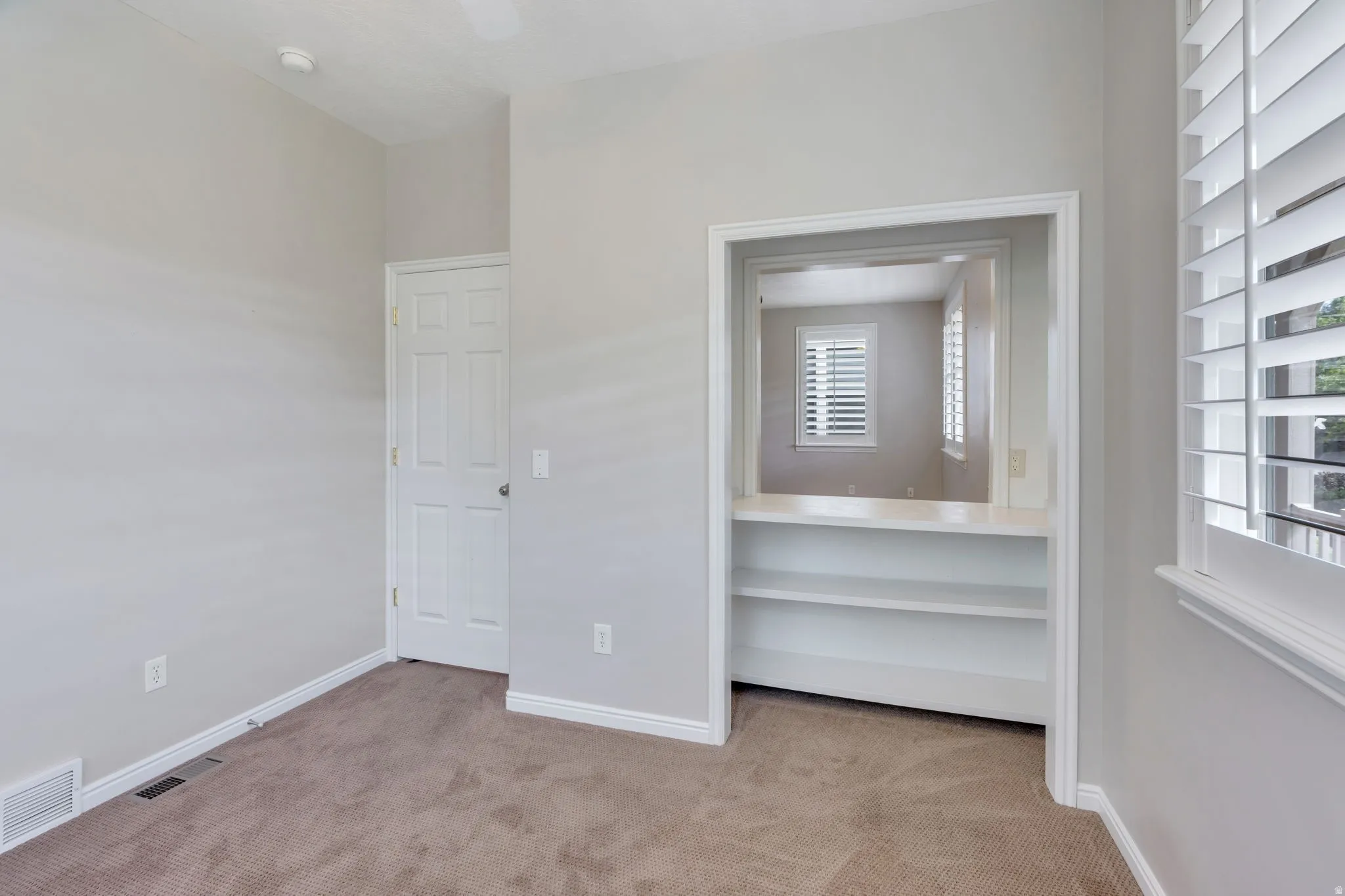 Neutral wall color, shuttered windows, built-in shelving, carpet flooring, and a white door