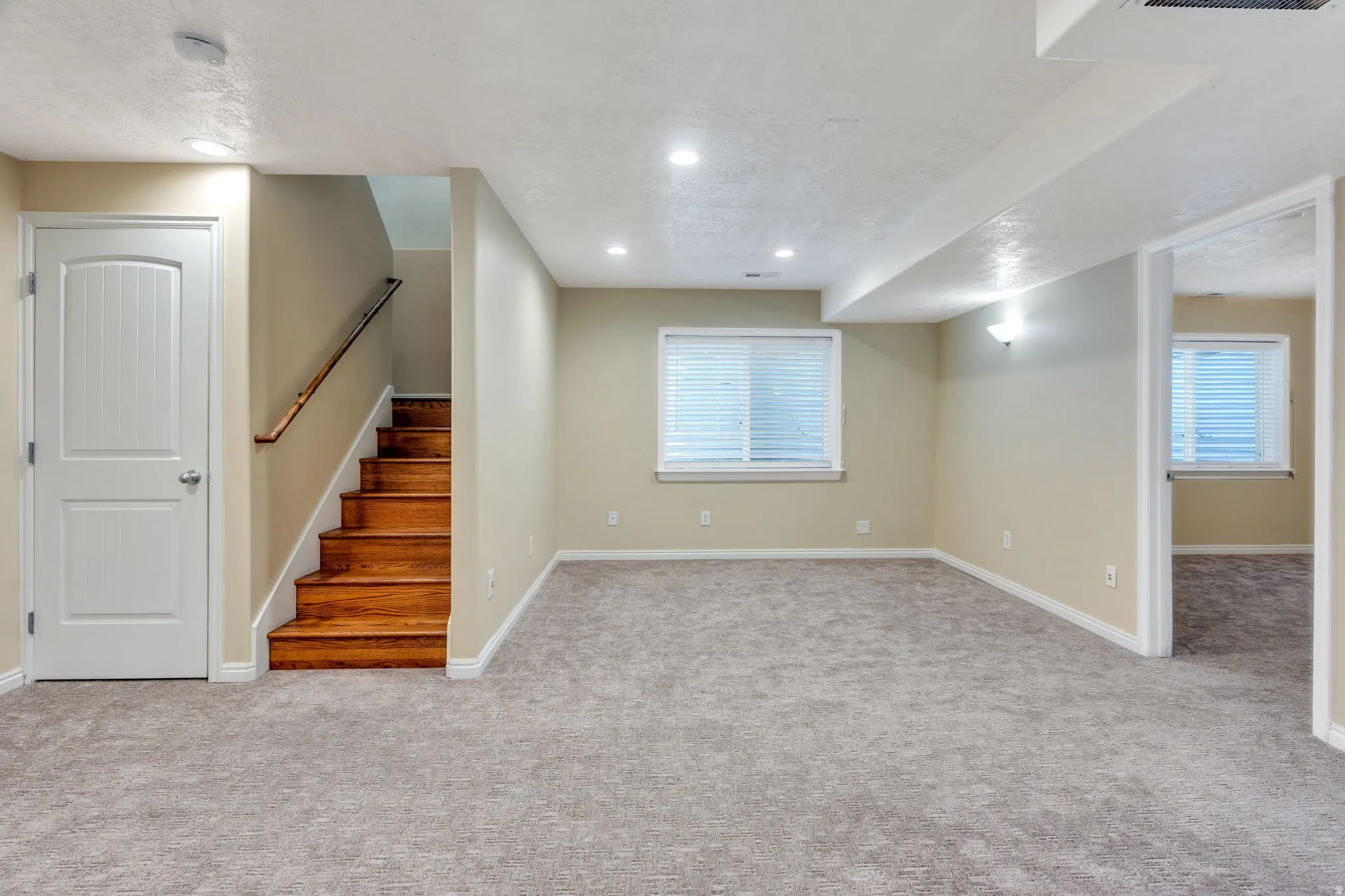 Spacious carpeted room featuring recessed lighting, multiple windows, a wood-tread staircase, and a paneled interior door