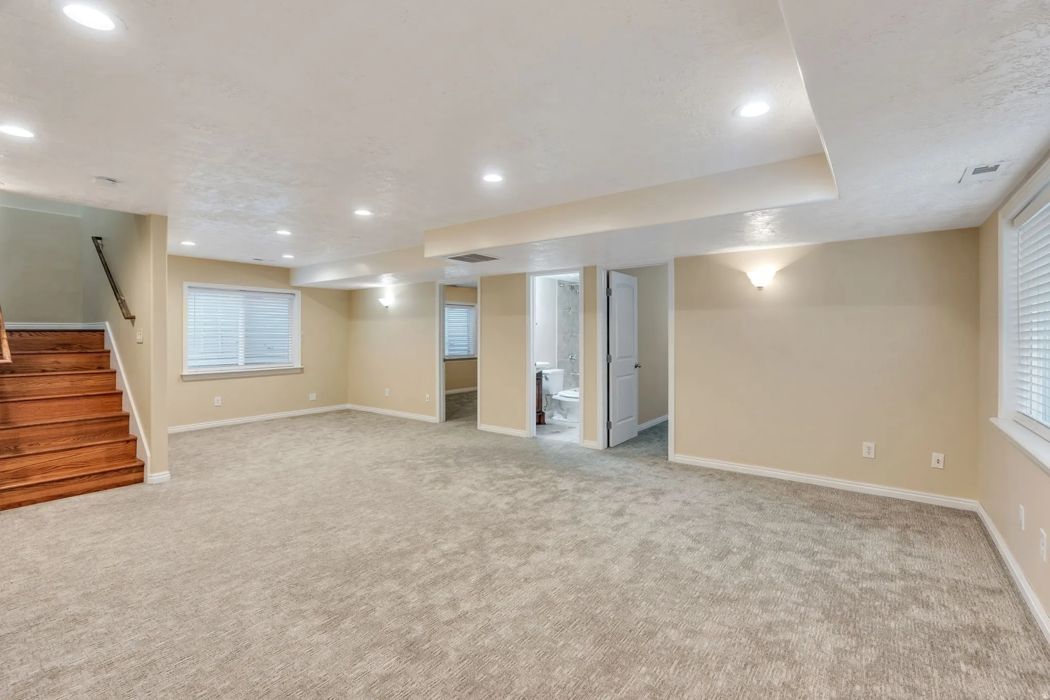 Spacious carpeted room featuring recessed lighting, a wood-finish staircase with a dark handrail, and multiple windows with blinds