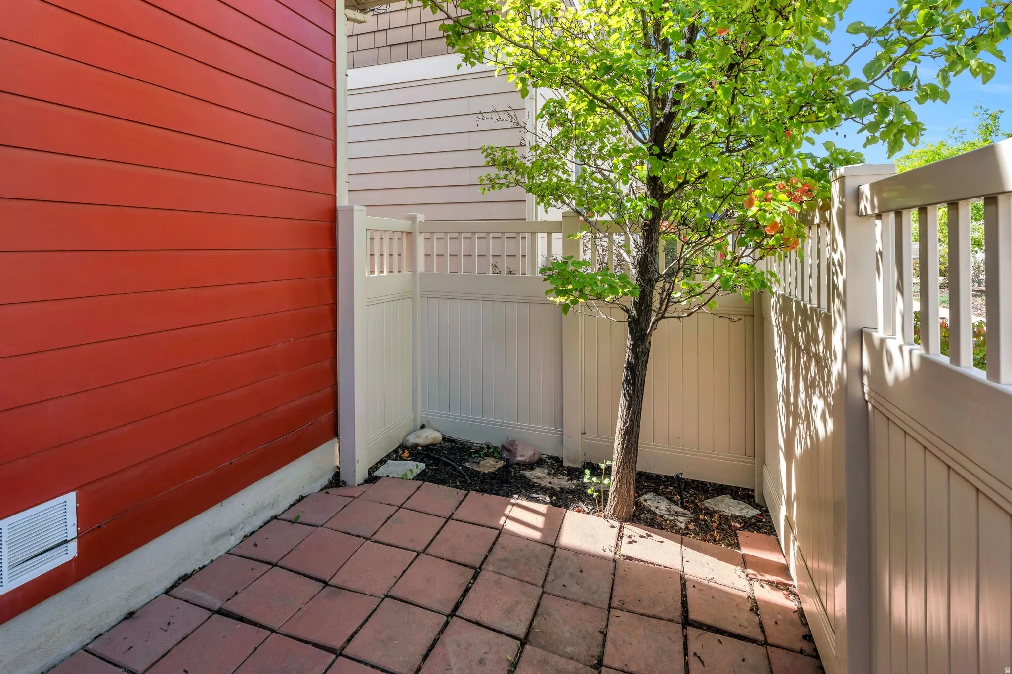 Paved patio area, contrasting red and beige siding, white privacy fencing, and a small tree
