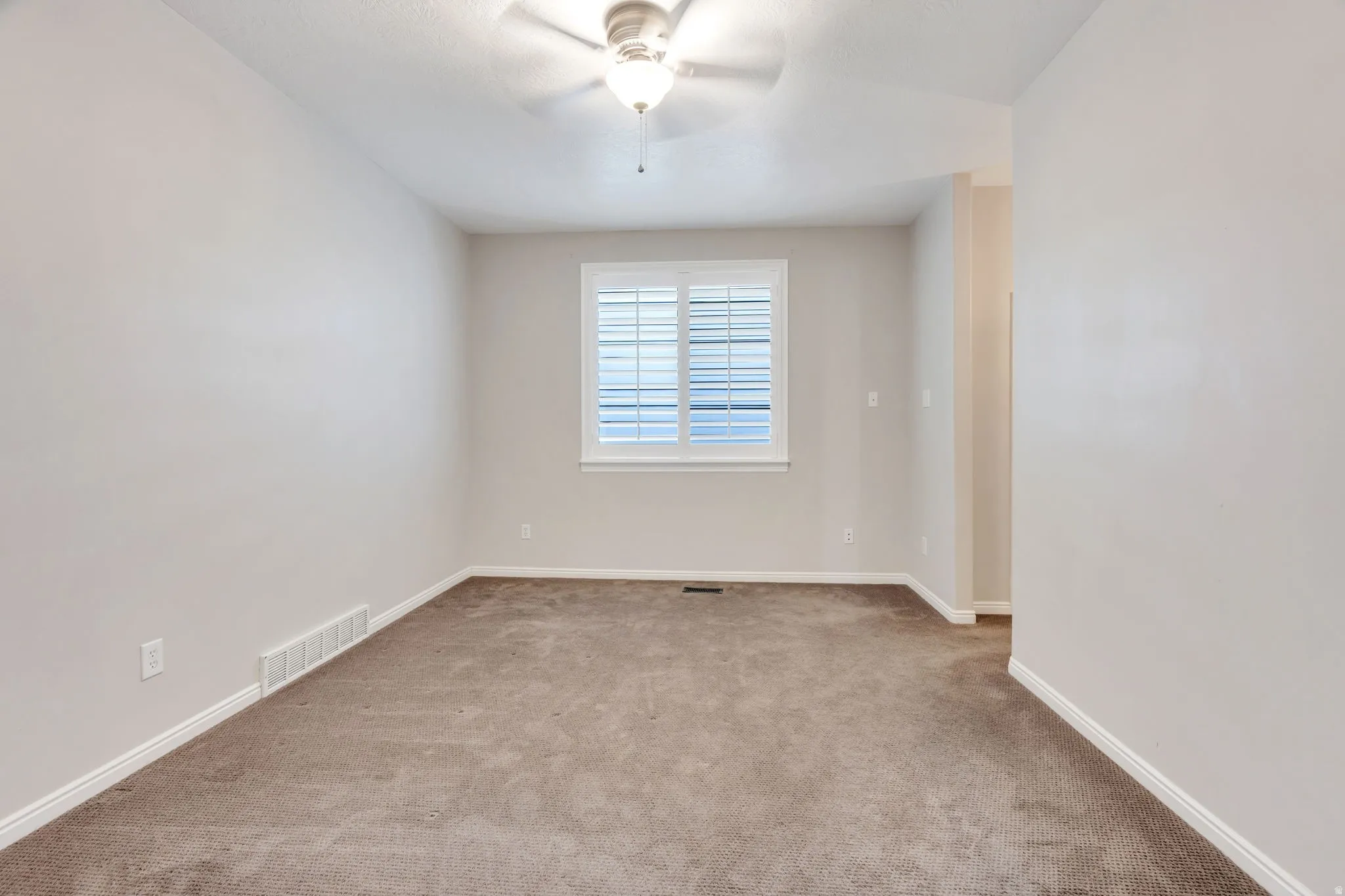 Neutral tone wall paint, textured ceiling, ceiling fan with integrated lighting, window with plantation shutters, and carpet flooring