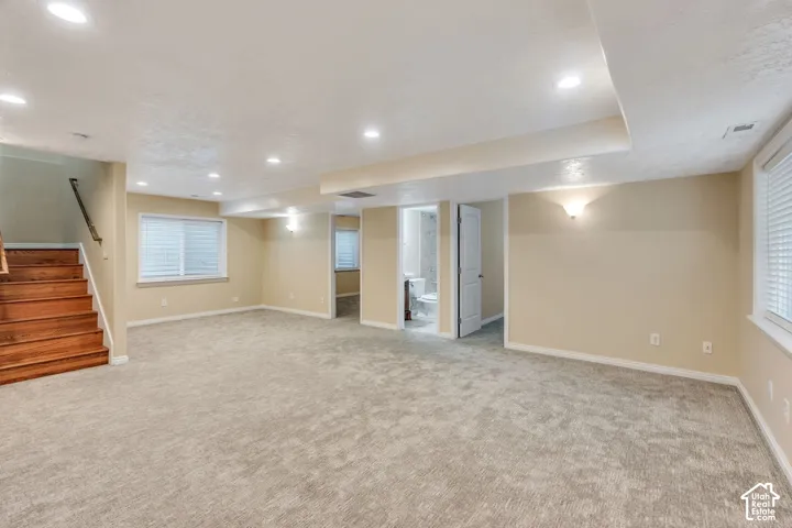 Spacious carpeted room featuring recessed lighting, a wood-finish staircase with a dark handrail, and multiple windows with blinds