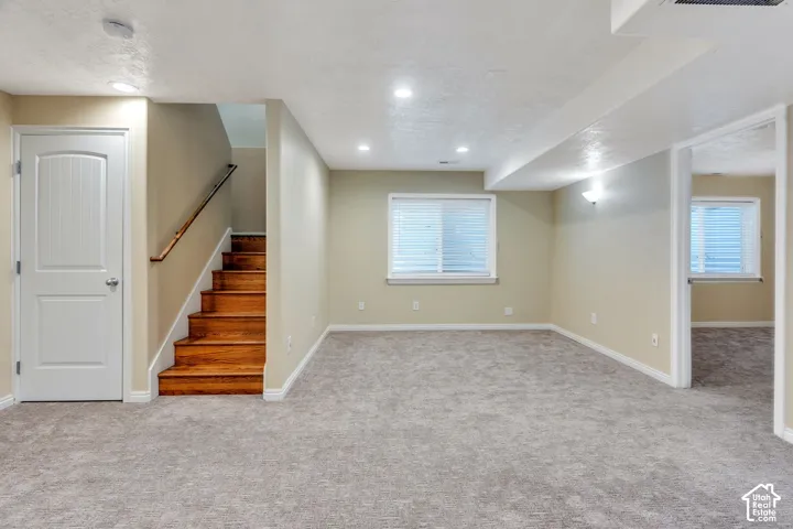 Spacious carpeted room featuring recessed lighting, multiple windows, a wood-tread staircase, and a paneled interior door