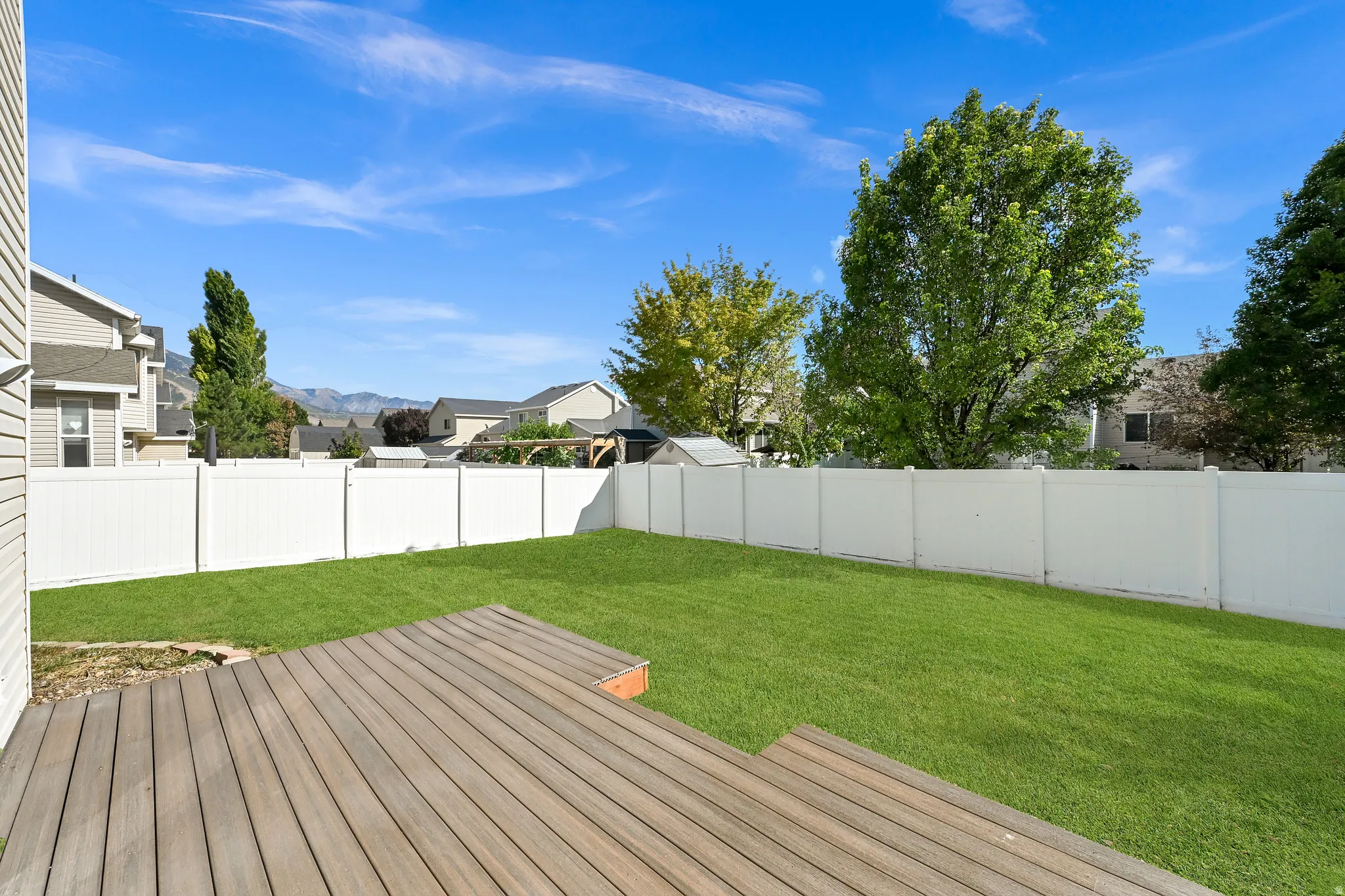 Expansive green lawn, multi-level wood-finish deck, white privacy fence, mature trees, and distant mountain backdrop