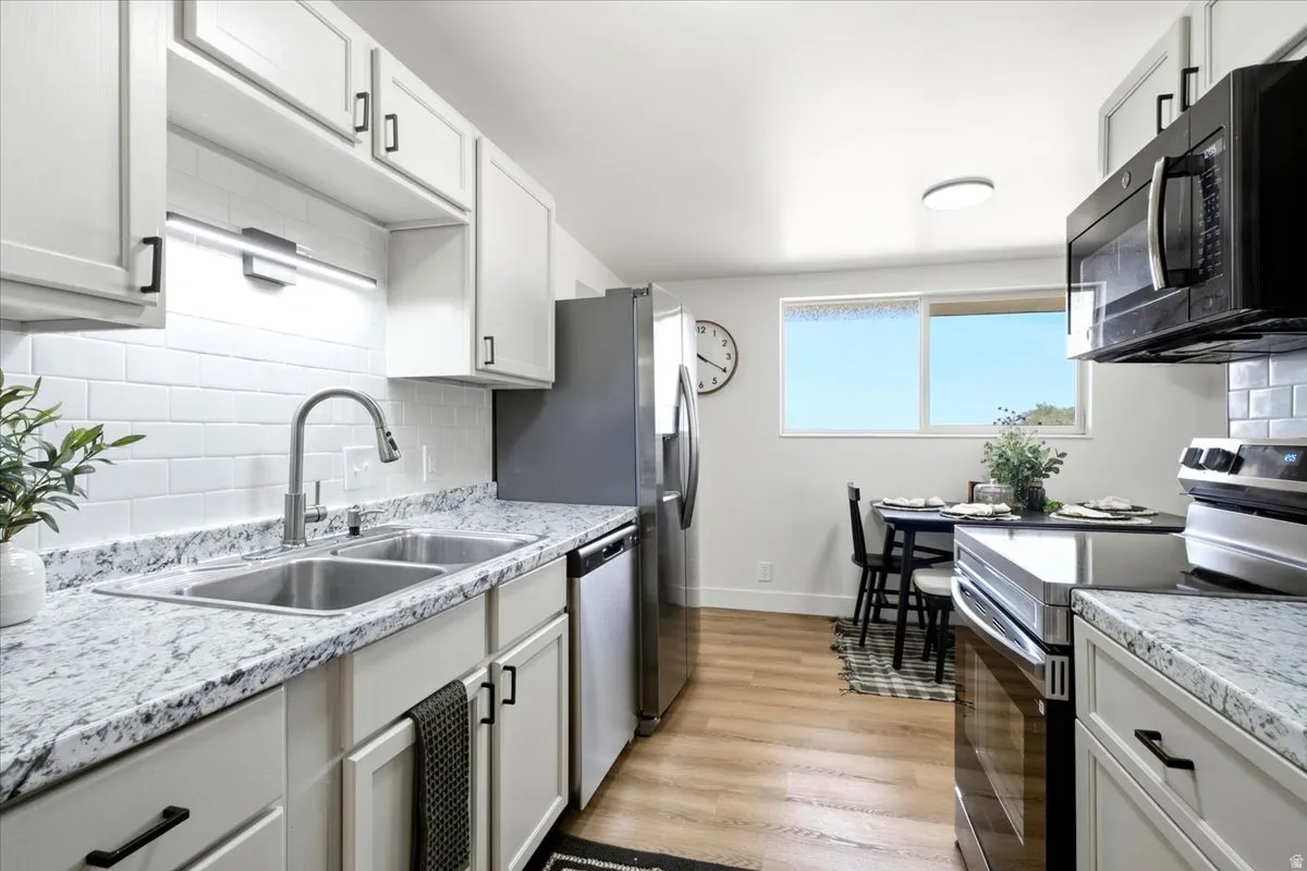 White shaker-style cabinetry with matte black hardware, dual stainless steel basin sink with a high-arc faucet, subway tile backsplash, and light-tone wood-finish flooring