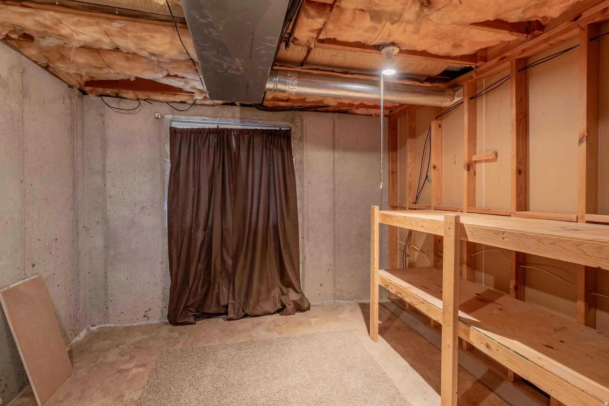 Unfinished basement space featuring exposed concrete walls, wood framing, utility lighting, and built-in wood shelving