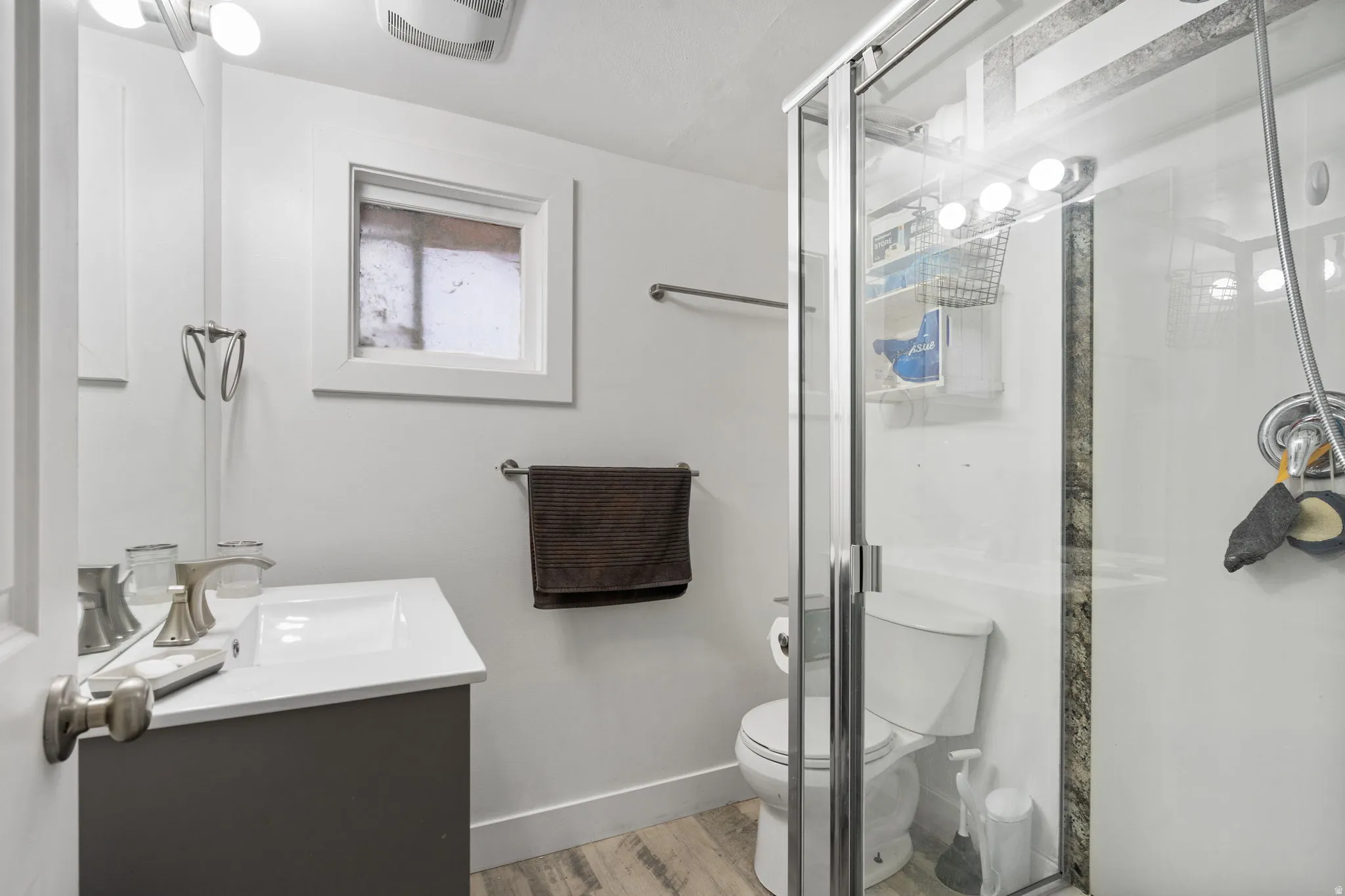 Glass-enclosed shower, single vanity with integrated sink, wood-finish flooring, and a compact window