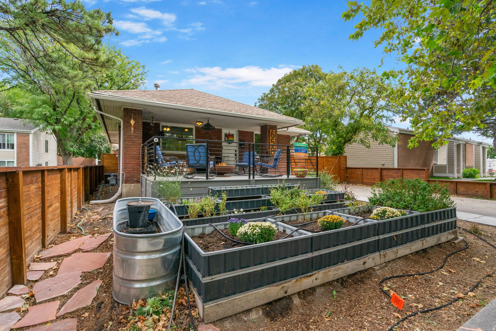 Elevated deck with metal railings, tiered planter boxes, stone pathway, wood fencing, and mature trees