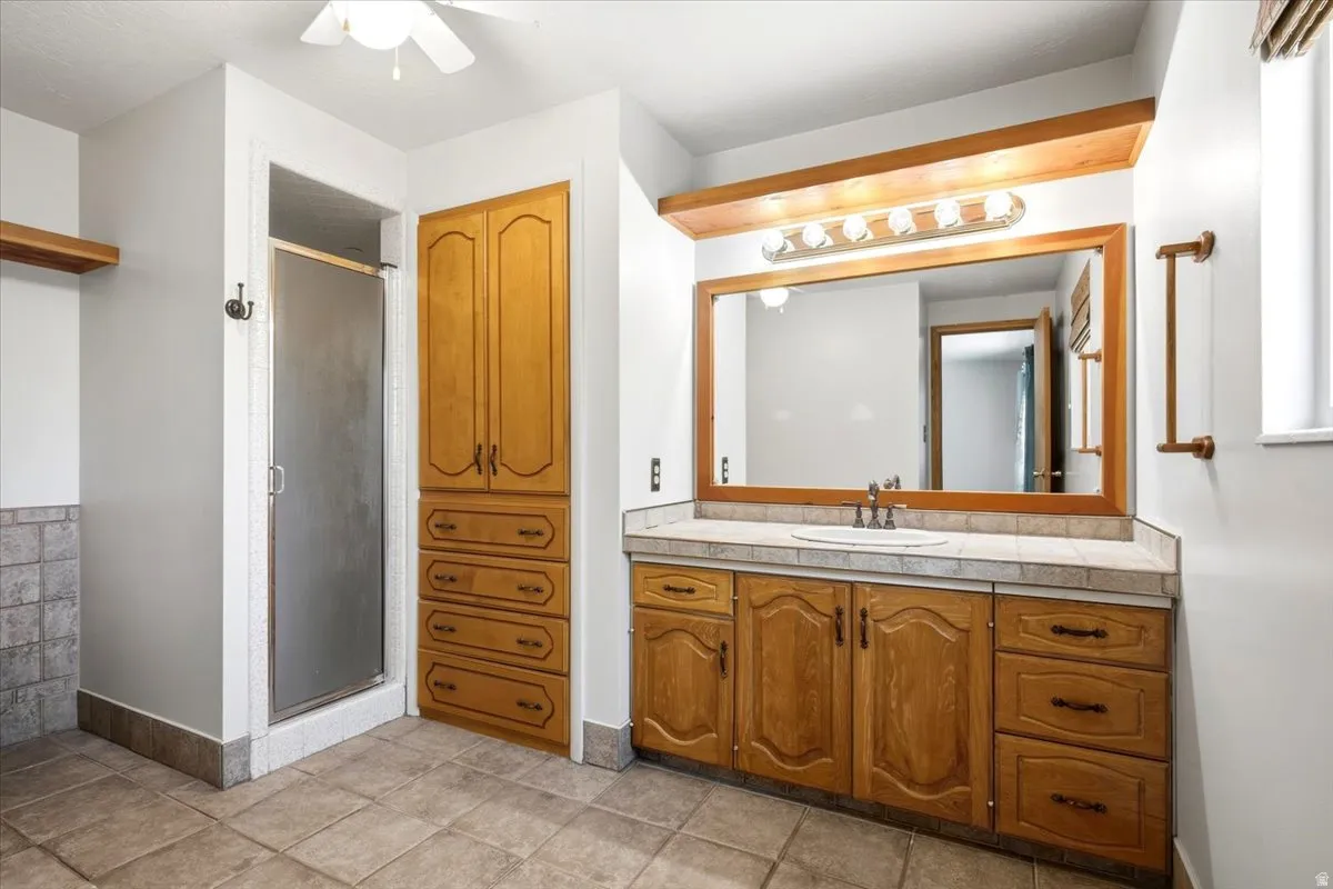 Tile flooring, wood-finish vanity with integrated sink, tile countertop, large framed mirror, and a standing shower with frosted glass