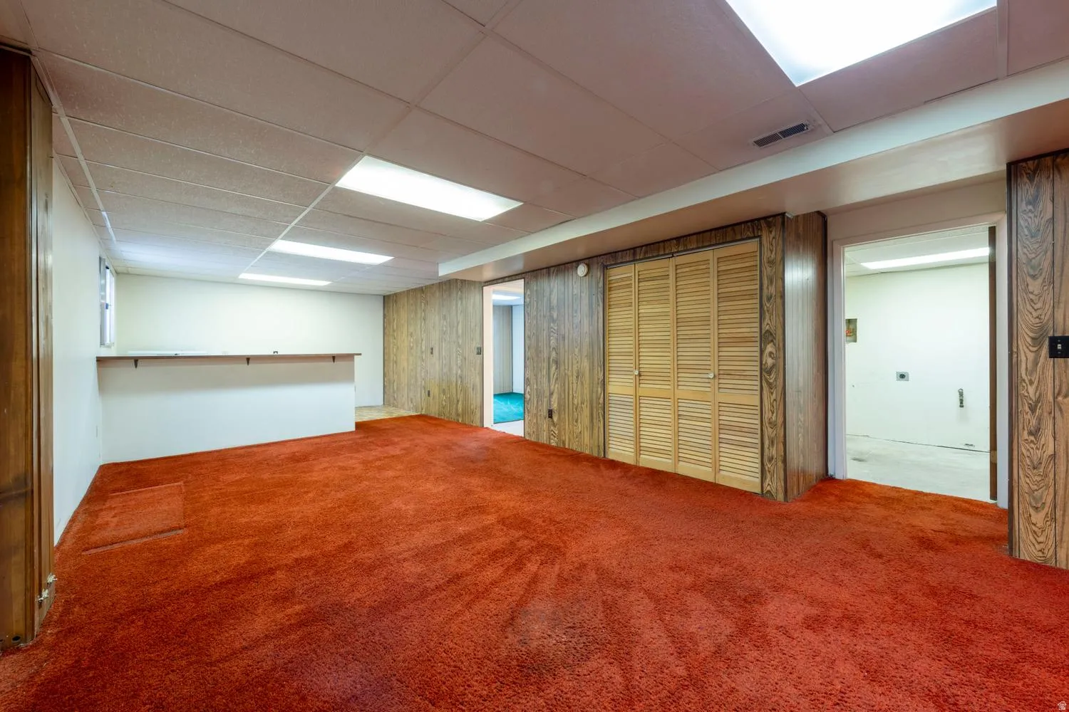 Finished room featuring broadloom carpeting, wood-paneled walls, a drop ceiling with integrated lighting, and louvered bifold closet doors