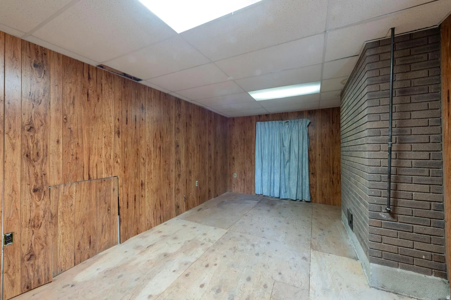 Wood-finish wall paneling, drop ceiling with integrated lighting, exposed brick wall feature, and unfinished subfloor