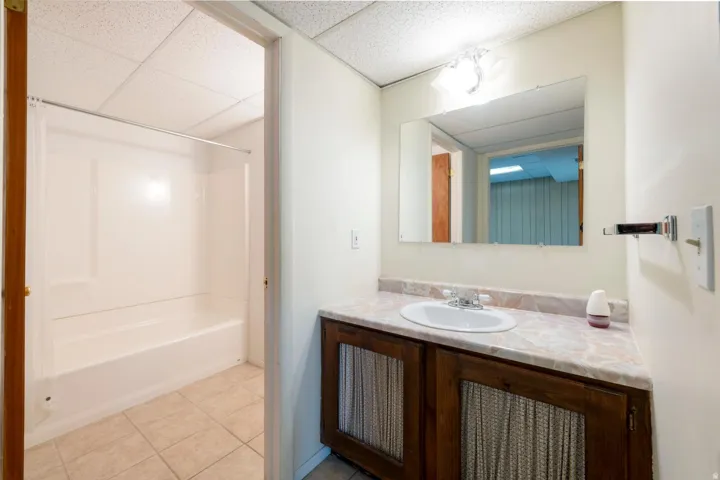 White integrated shower and tub unit, tile flooring, vanity with integrated sink, wood-finish cabinetry, and wall-mounted mirror