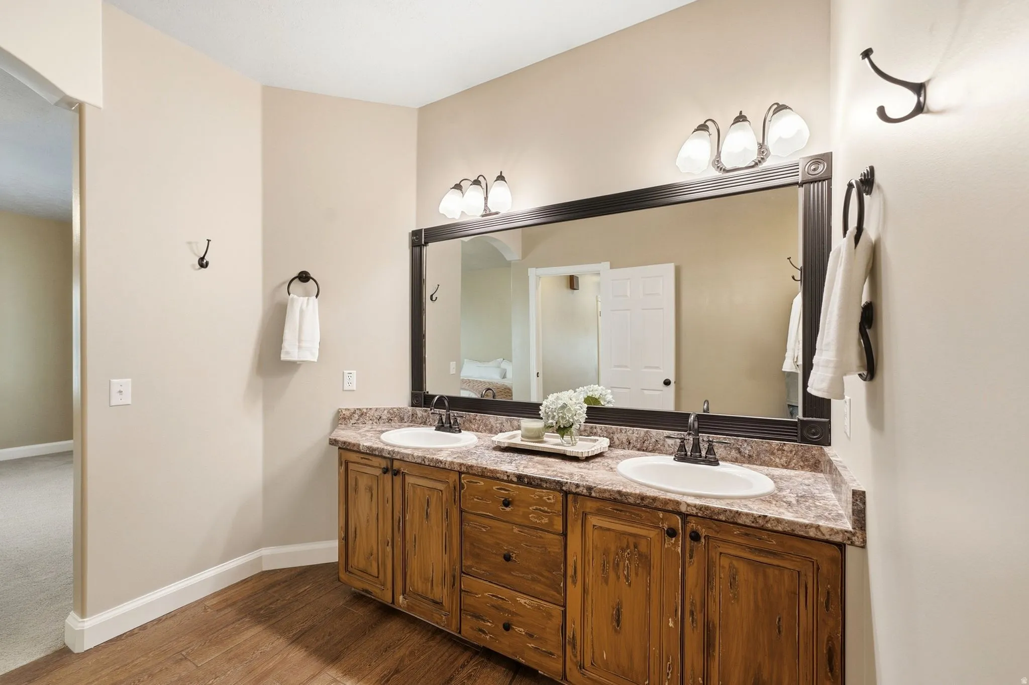 Dual vanity with integrated sinks, wood-finish cabinetry, a large framed mirror, a three-light fixture, and wood-finish flooring