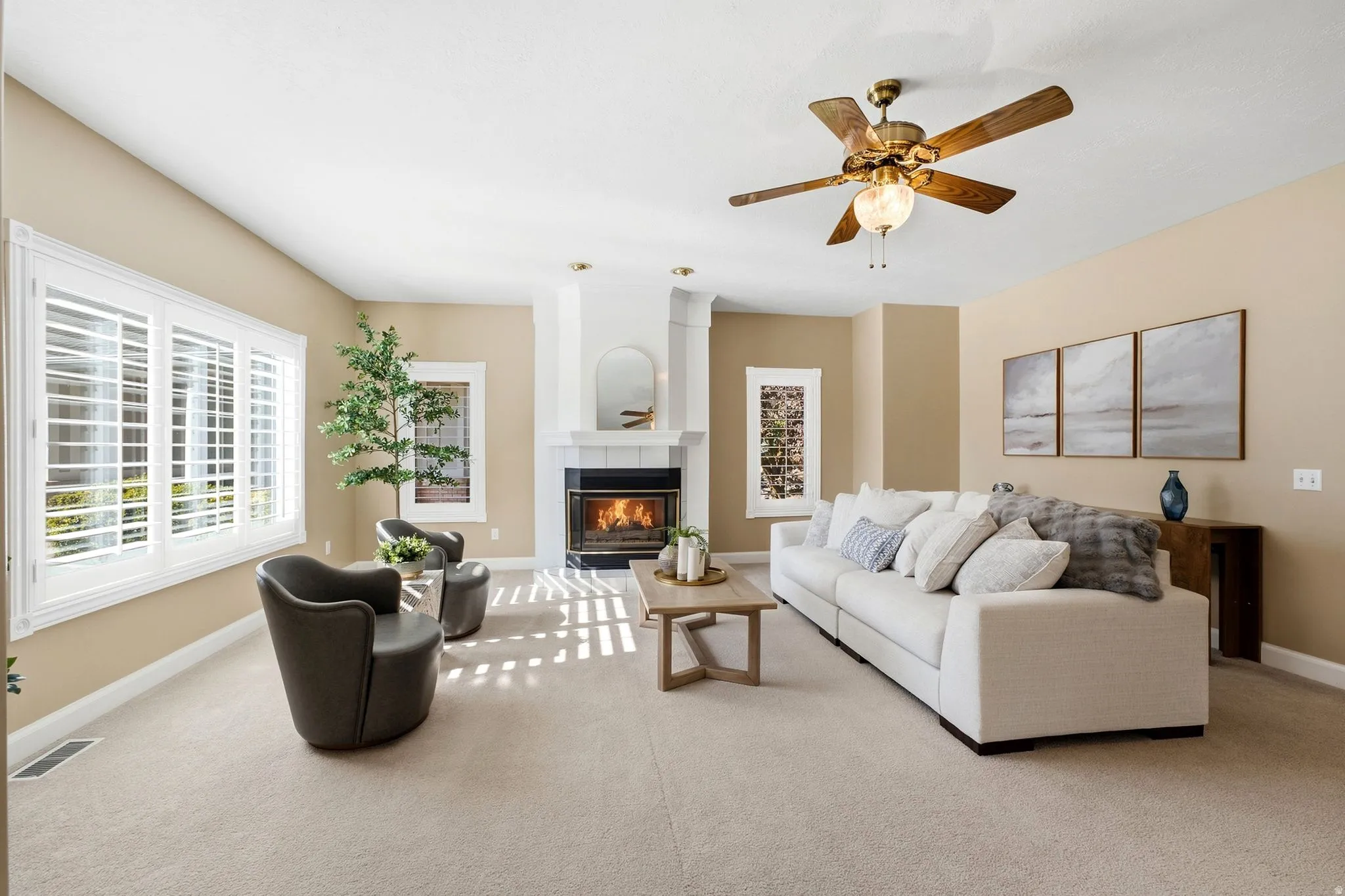 Spacious living area featuring a fireplace with a white mantel, ceiling fan, carpet flooring, and large windows with white shutters