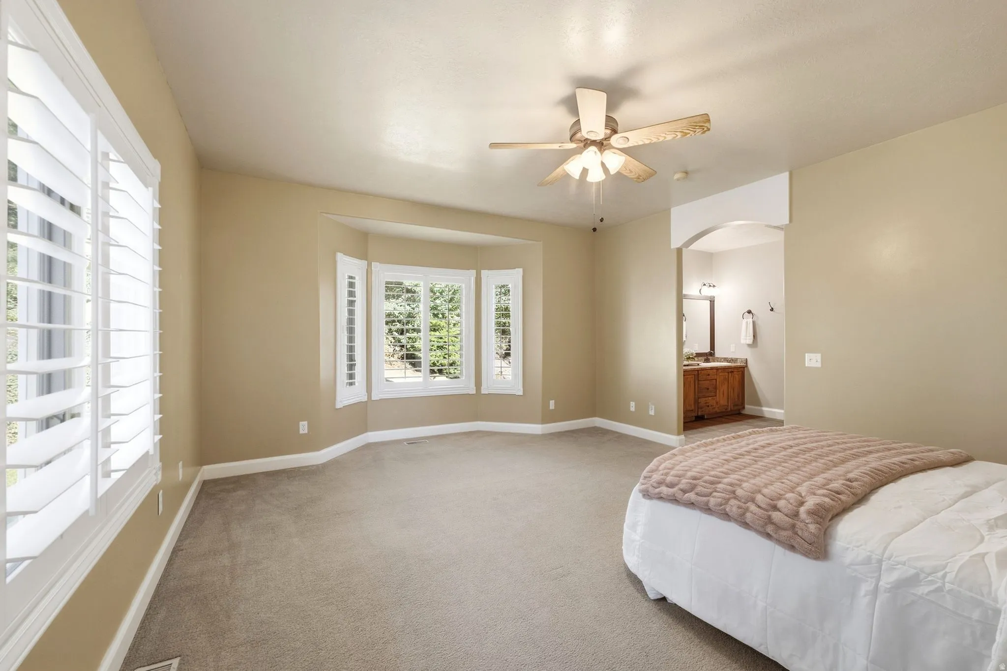 Carpeted room featuring a bay window with plantation shutters, ceiling fan, and arched doorway
