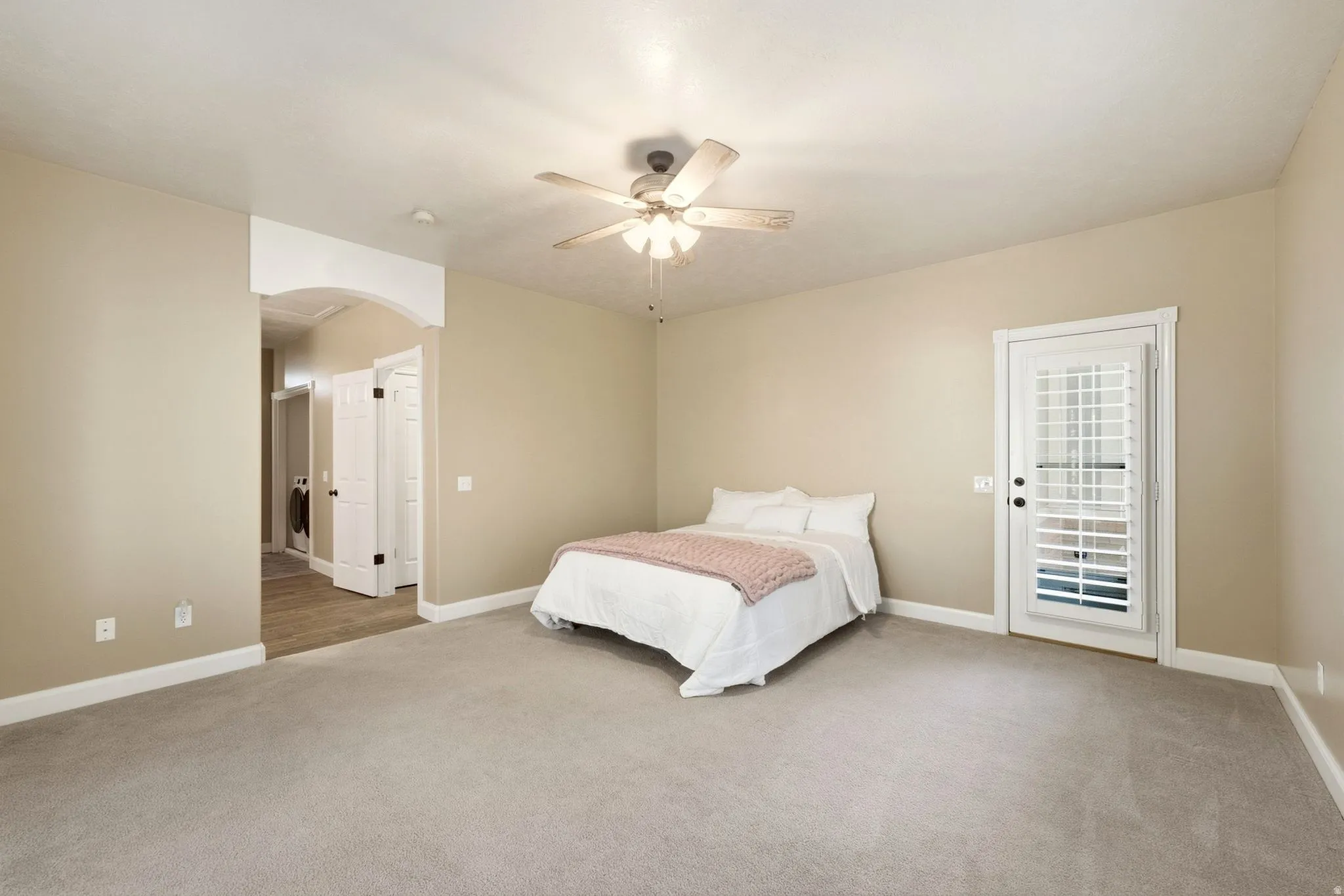 Spacious room featuring neutral carpeting, a ceiling fan, a private exterior door with louvered blinds, and an arched entry