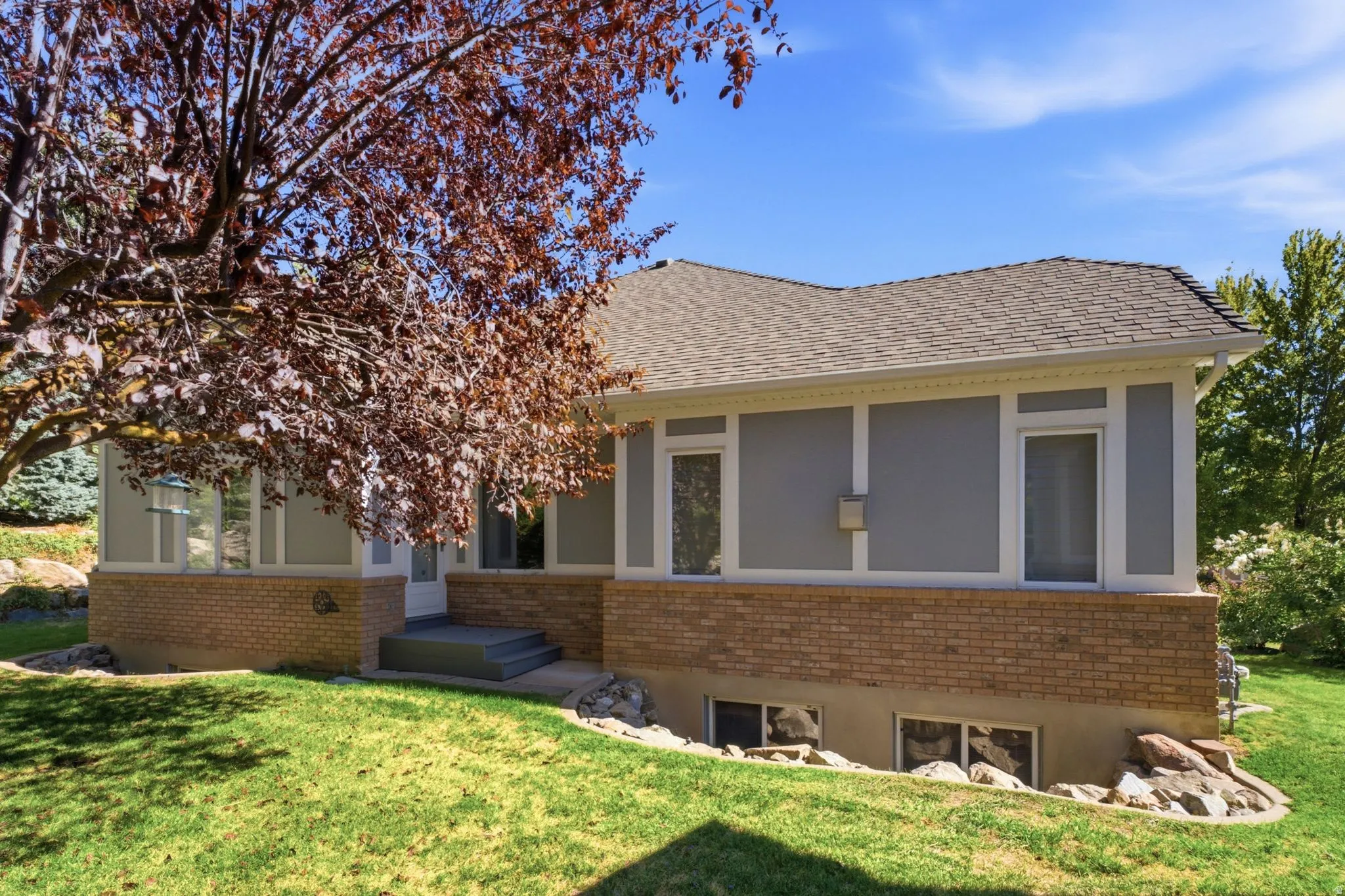 Shingled roof, light gray siding, brick facade, white trim, and a mature tree