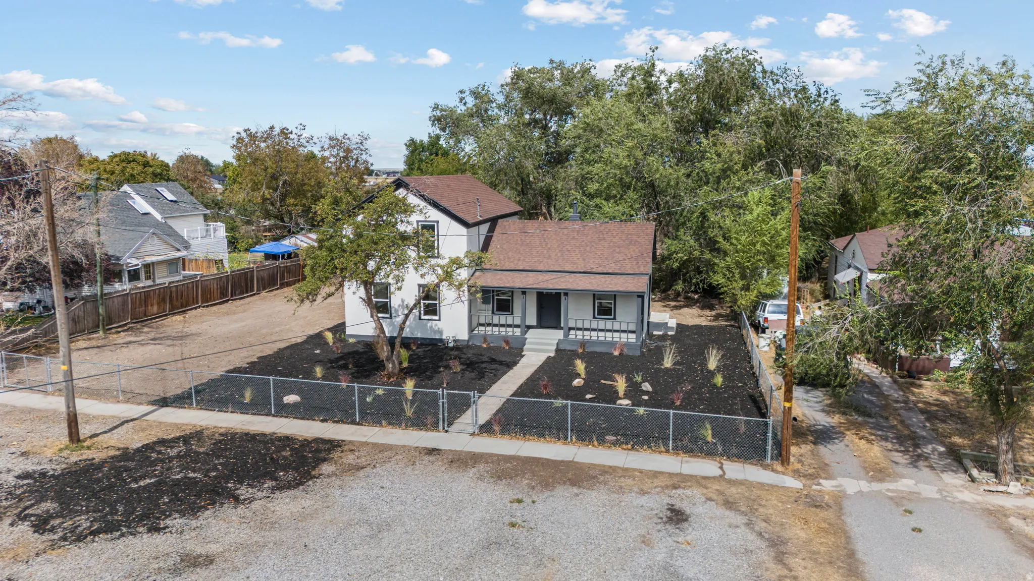 Two-story exterior with white siding and dark trim, featuring a covered porch, a newly landscaped front yard with black mulch and young plantings, and a chain-link fence