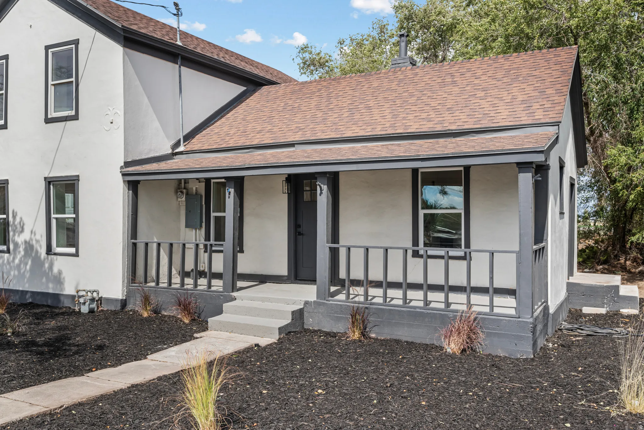 Front porch with dark railing and columns, light stucco exterior, dark trim windows, and a shingle roof
