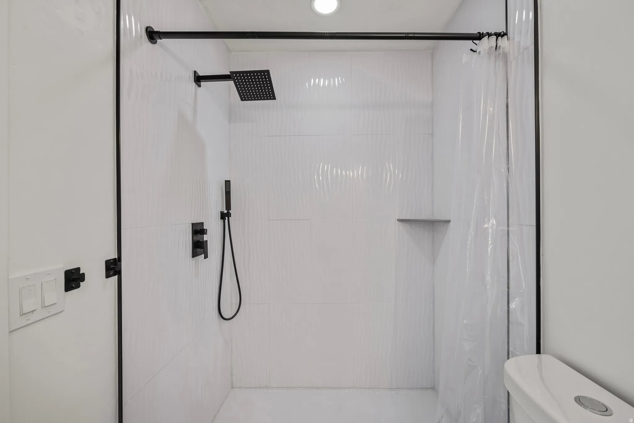 Walk-in shower featuring textured white tile, a matte black rainfall showerhead, and a built-in corner shelf
