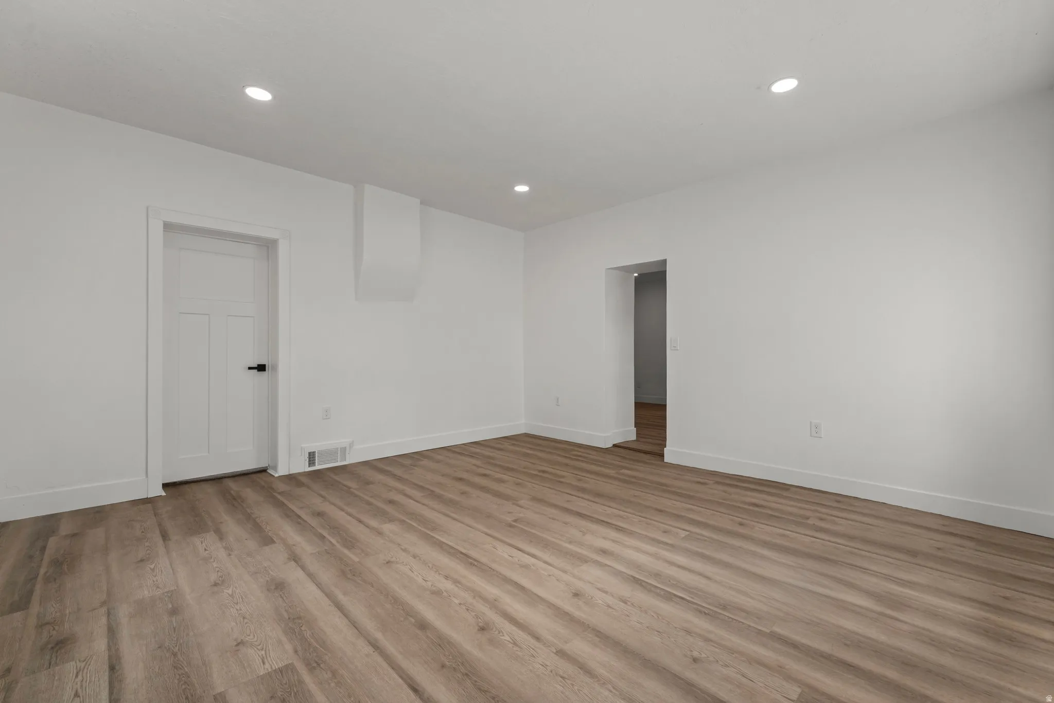 Recessed lighting, wood-finish flooring, white walls, and a white paneled door with black hardware