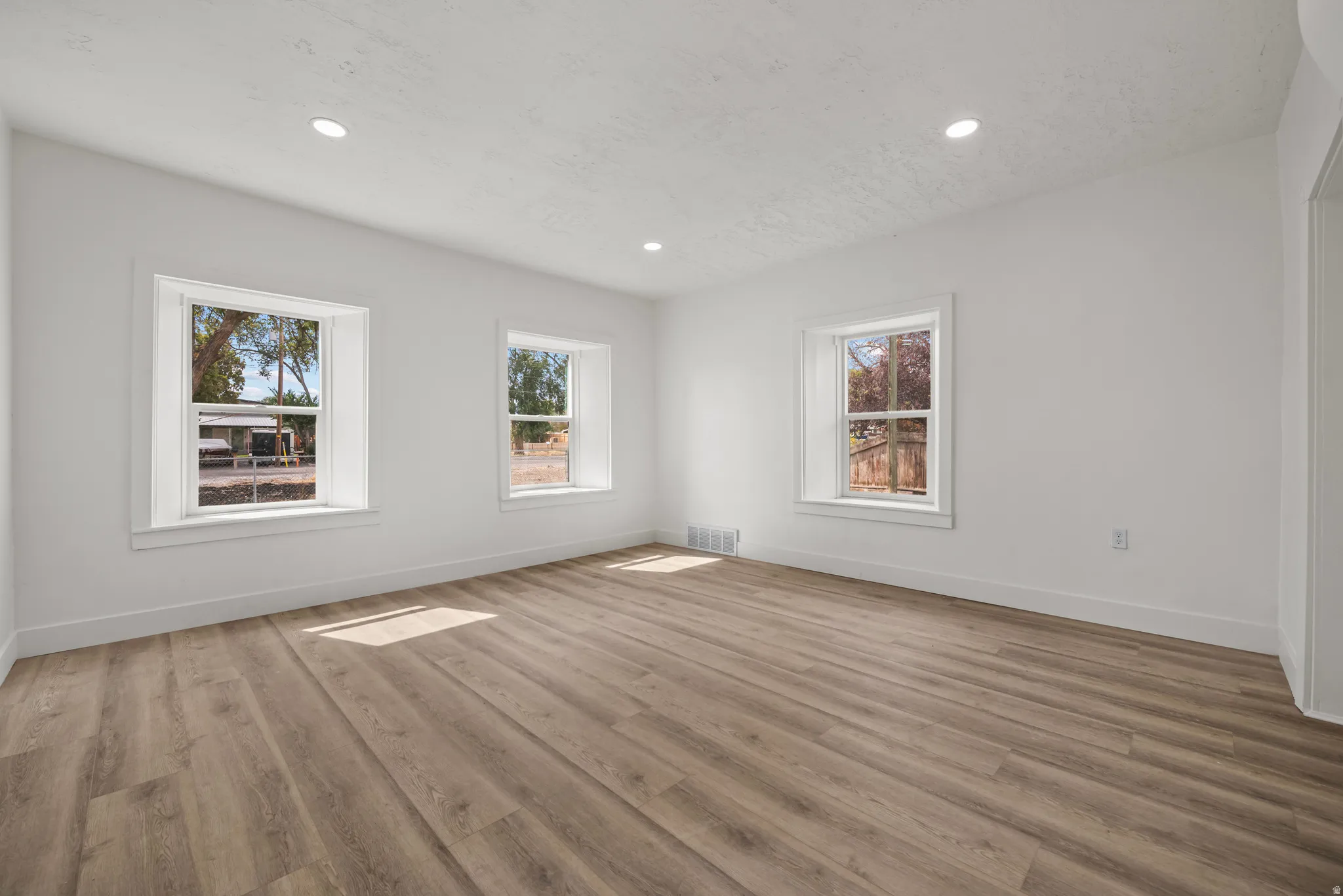 Recessed lighting illuminates the space, featuring three windows with white trim, wood-finish flooring, and crisp white walls