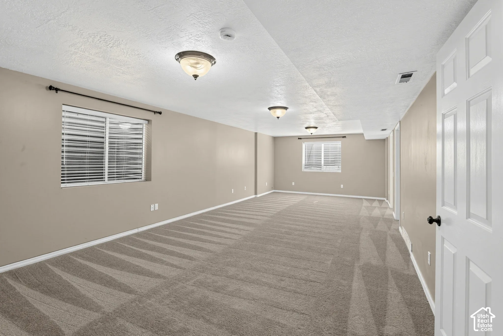 Spacious room with neutral-toned walls, patterned carpet, and multiple windows with blinds
