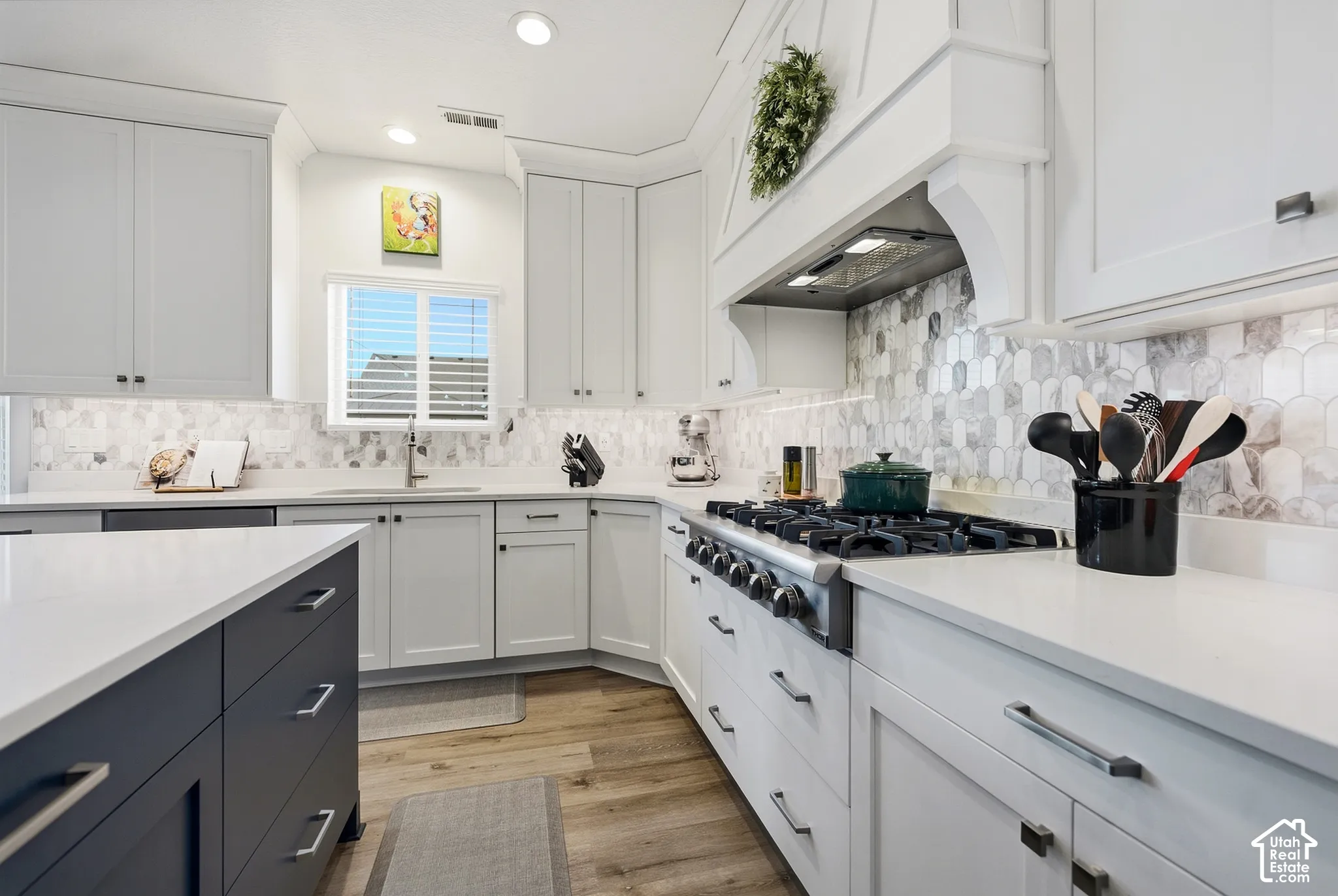 White cabinetry with under-cabinet lighting, a built-in range hood, a stainless-steel gas range, light-toned countertops, and a patterned backsplash