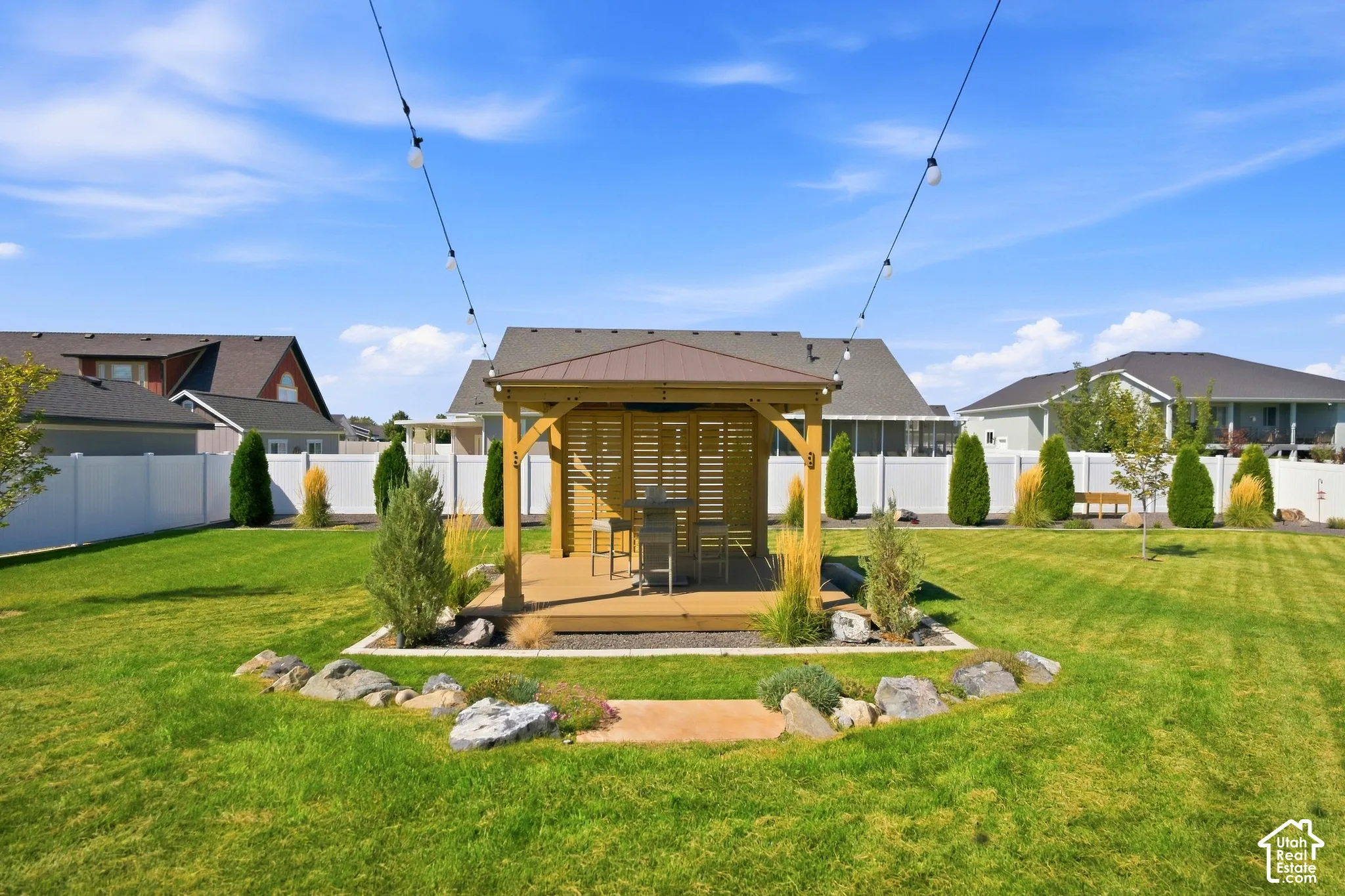 Expansive backyard featuring a central gazebo with a metal roof and slatted wood-finish walls, a surrounding wood-finish deck, a manicured lawn, and perimeter white privacy fencing