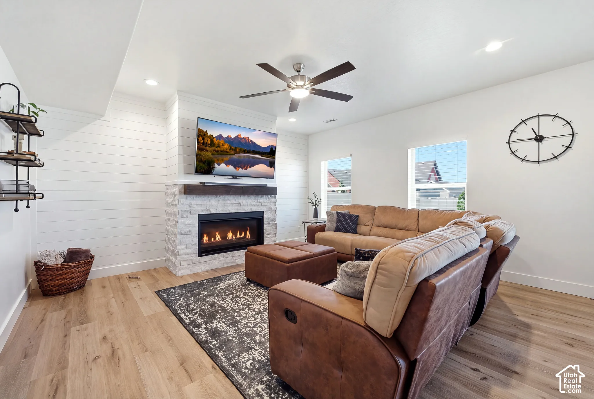 Living area featuring a stone-clad fireplace with a dark wood mantel, wood-finish flooring, white shiplap accent wall, and a ceiling fan with integrated lighting