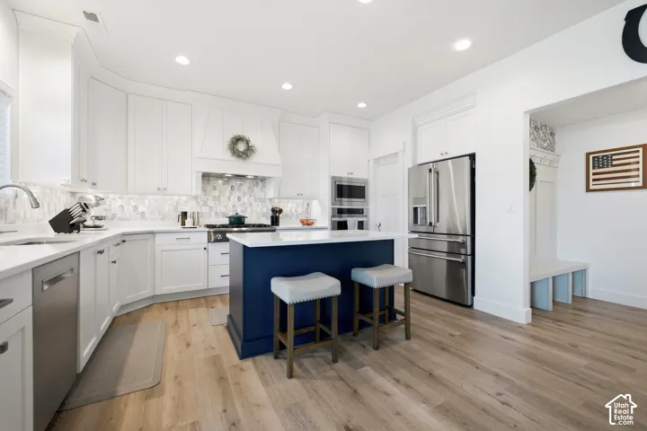 White cabinetry with undermount lighting, dark blue kitchen island, stainless steel appliances, patterned backsplash, and wood-finish flooring