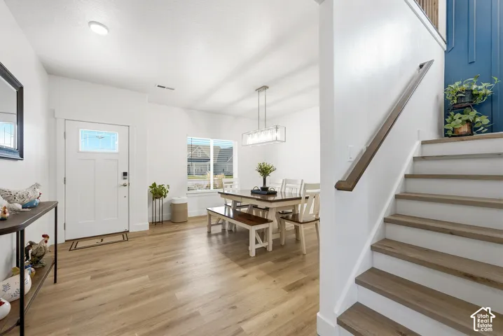 Wood-finish flooring throughout, white paneled entry door with decorative glass, large window with white trim, contemporary linear chandelier, and a staircase with wood-finish treads and handrail