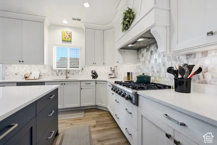 White cabinetry with under-cabinet lighting, a built-in range hood, a stainless-steel gas range, light-toned countertops, and a patterned backsplash
