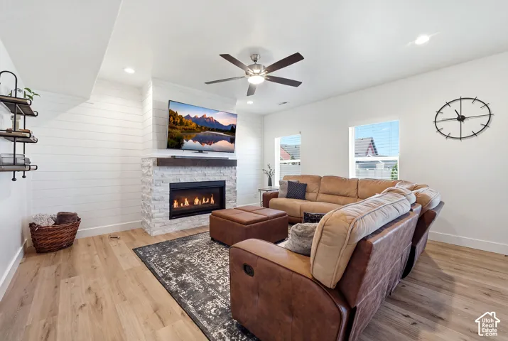 Living area featuring a stone-clad fireplace with a dark wood mantel, wood-finish flooring, white shiplap accent wall, and a ceiling fan with integrated lighting