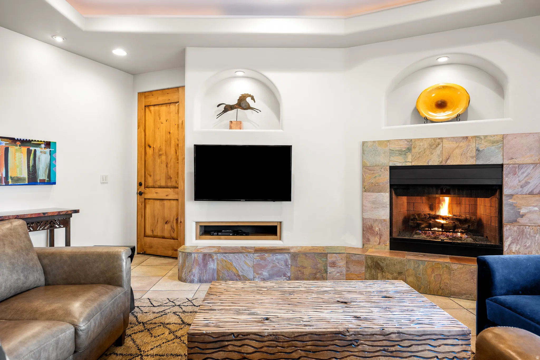 Corner fireplace with stone surround, built-in display niches, recessed ceiling lighting, wood-finish door, and tile flooring