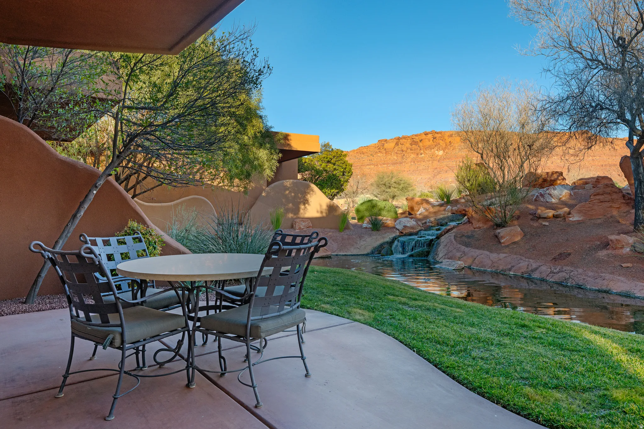 Paved patio area, a water feature with a waterfall, manicured lawn, and a natural desert backdrop with red rock formations