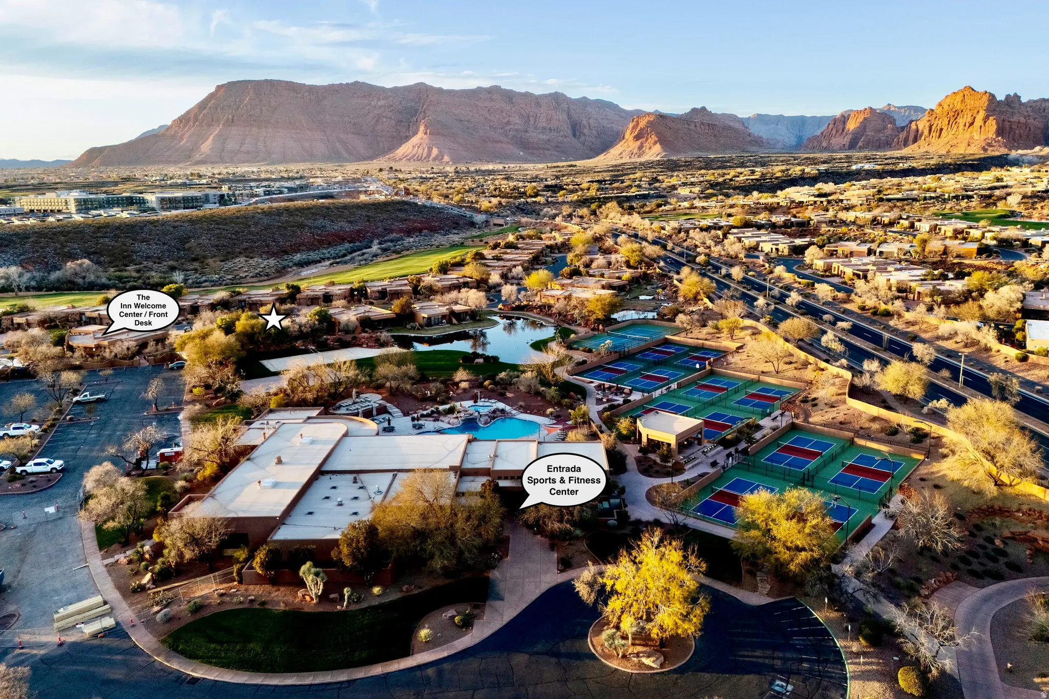 Expansive aerial panorama showcasing a large sports and fitness center, multiple tennis courts, a swimming pool complex, and a serene body of water, all set against a dramatic mountainous backdrop