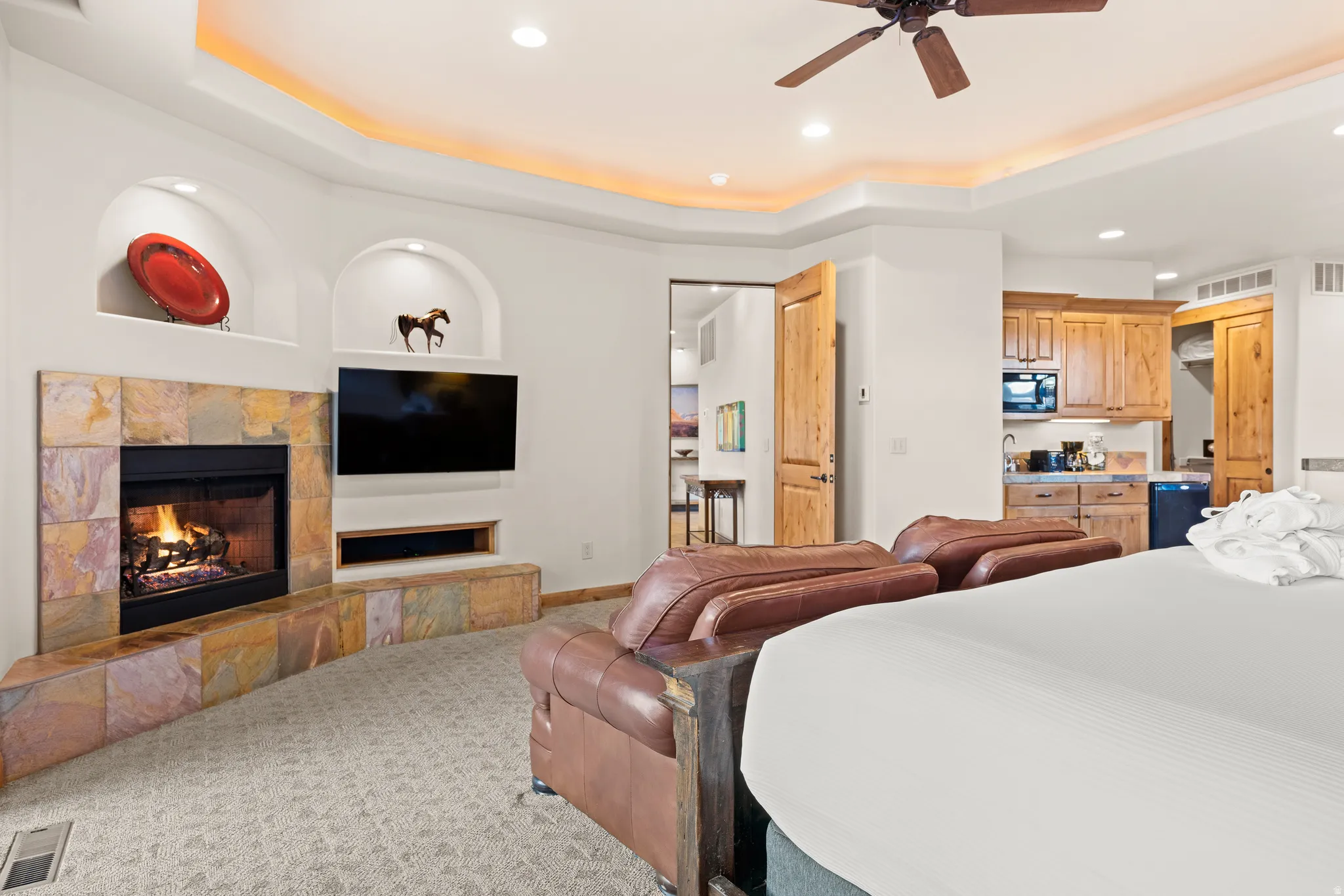Built-in fireplace with stone surround, recessed wall niches, tray ceiling with recessed lighting, integrated ceiling fan, and compact kitchenette with wood-finish cabinetry