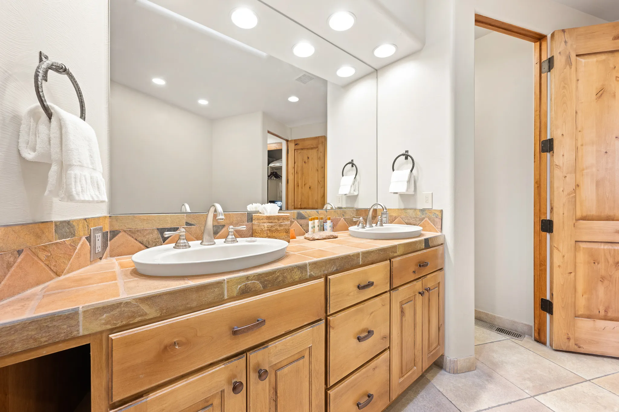Dual vanity with integrated sinks, wood cabinetry, tile countertop, and a mirrored wall with overhead lighting