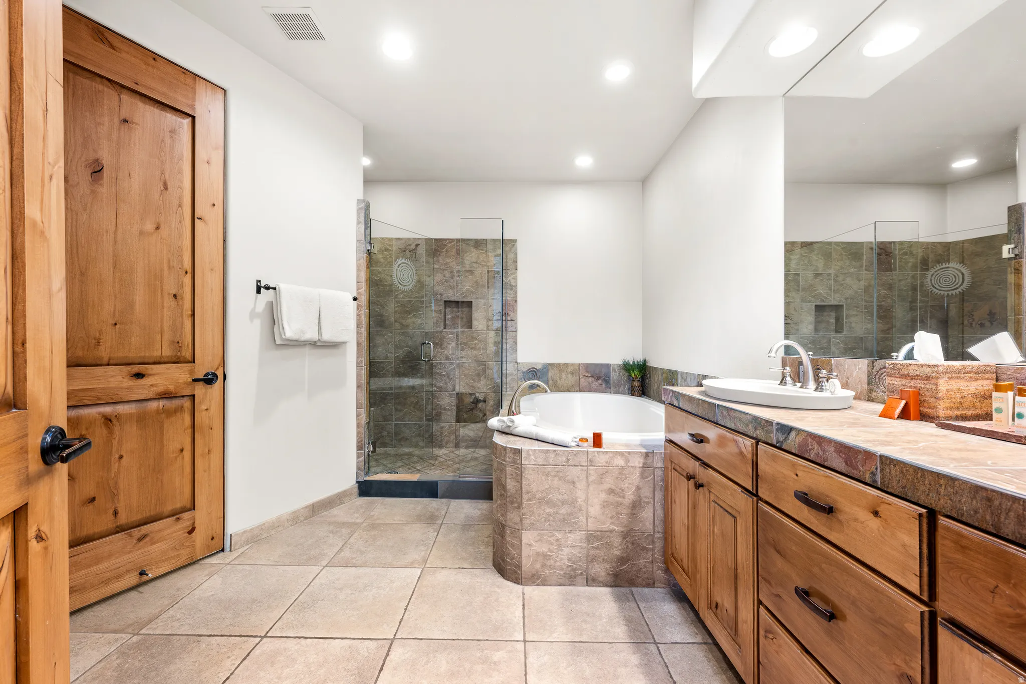 Recessed lighting illuminates the bathroom featuring a built-in tub, a glass-enclosed shower with tiled walls, and a wood-finish vanity with an undermount sink