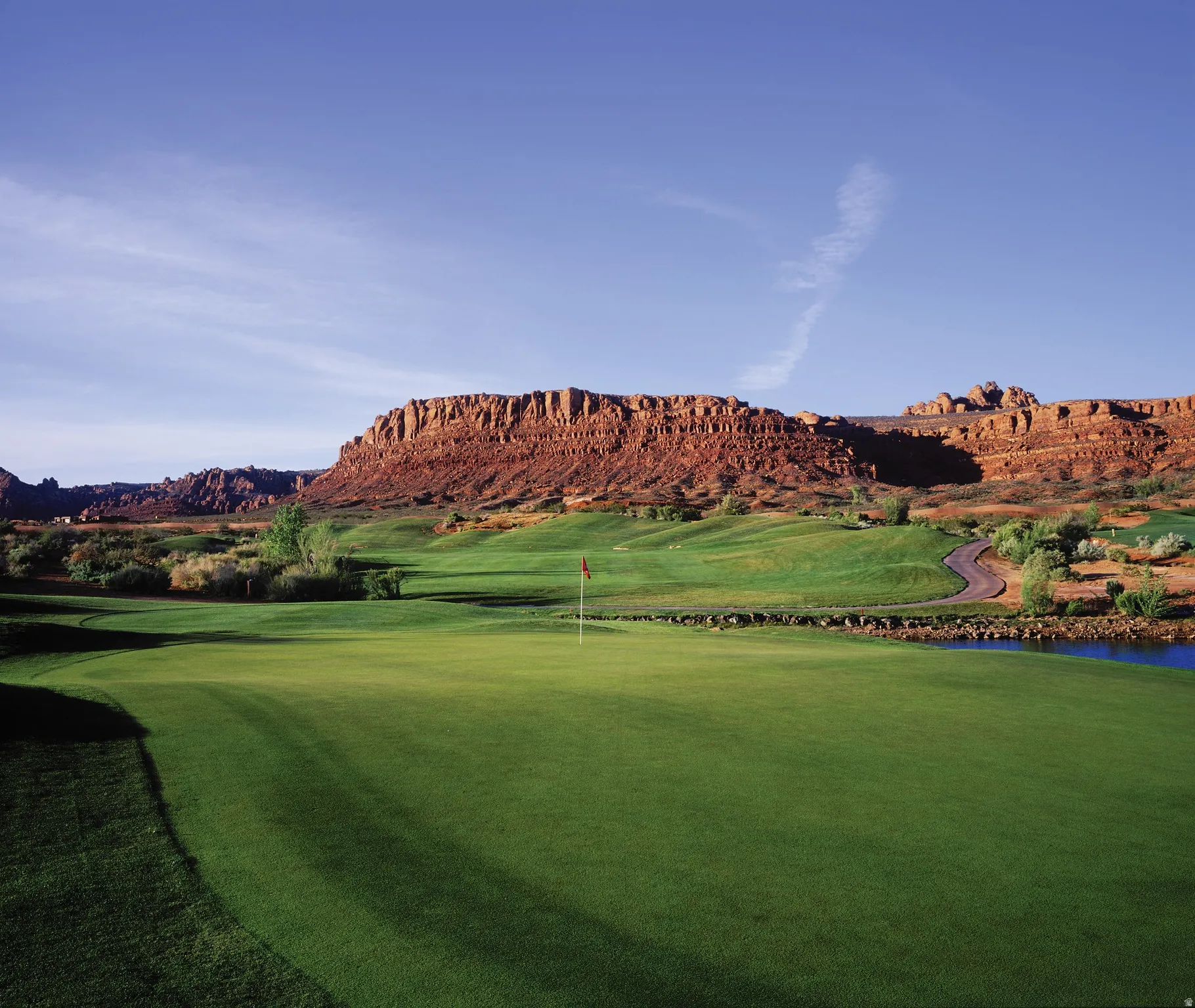 Expansive green with a red flag, surrounded by sculpted fairways and a paved path
