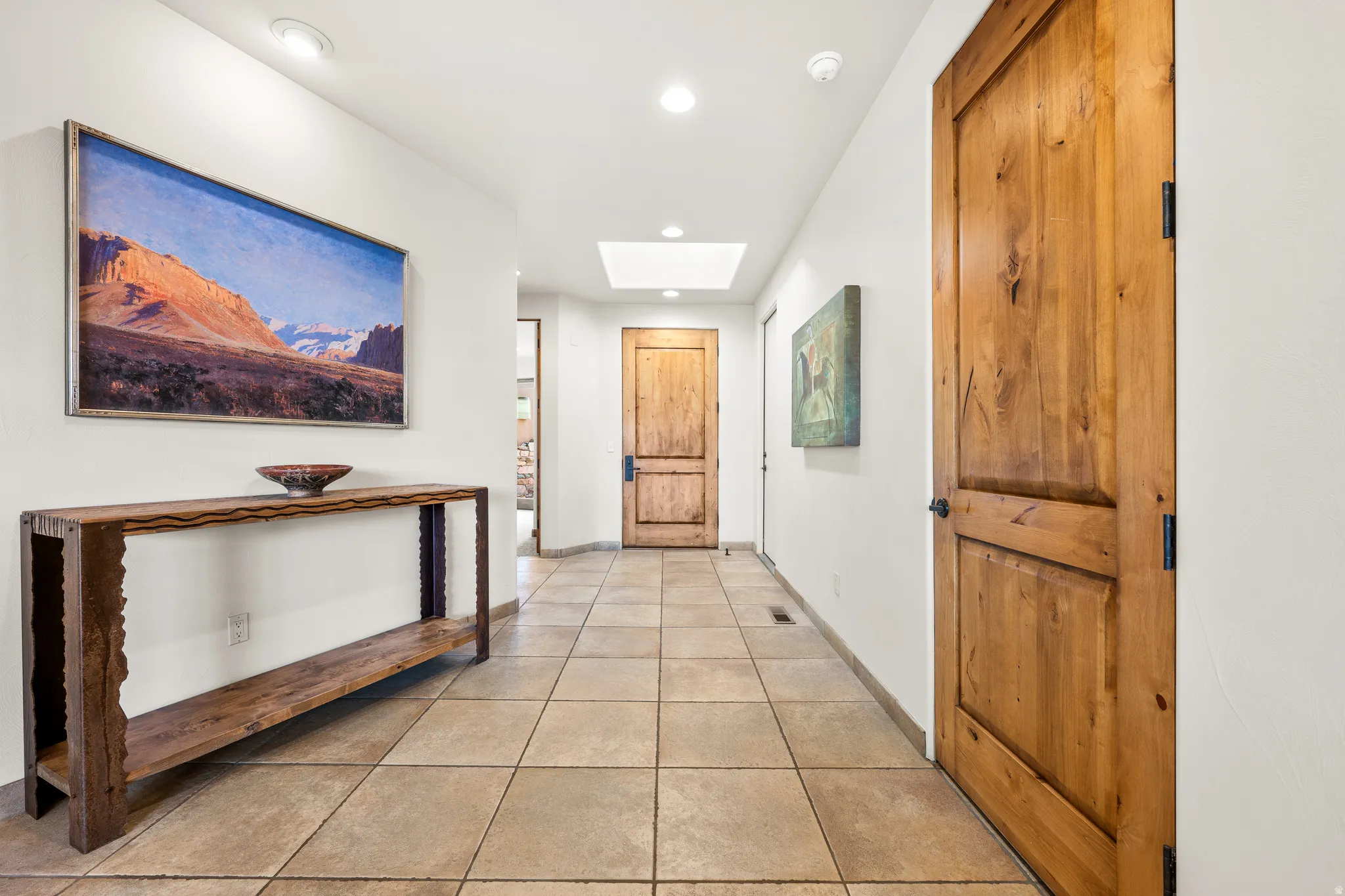 Recessed lighting, a skylight, and light-toned walls illuminate this hallway featuring large-format floor tiles and wood-panel doors