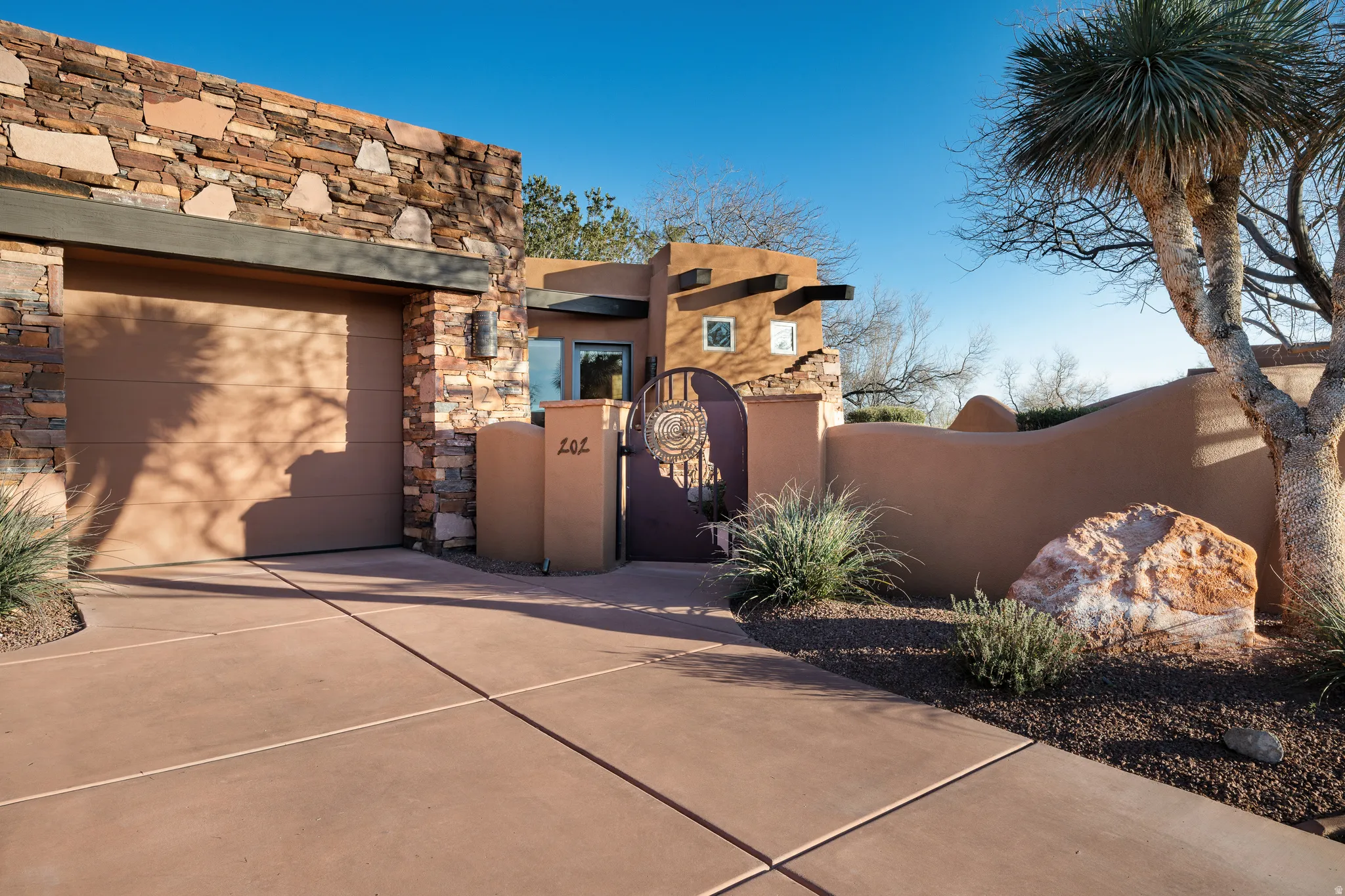 Stone veneer facade with an integrated garage, stucco exterior walls, wrought iron gate, and desert landscaping