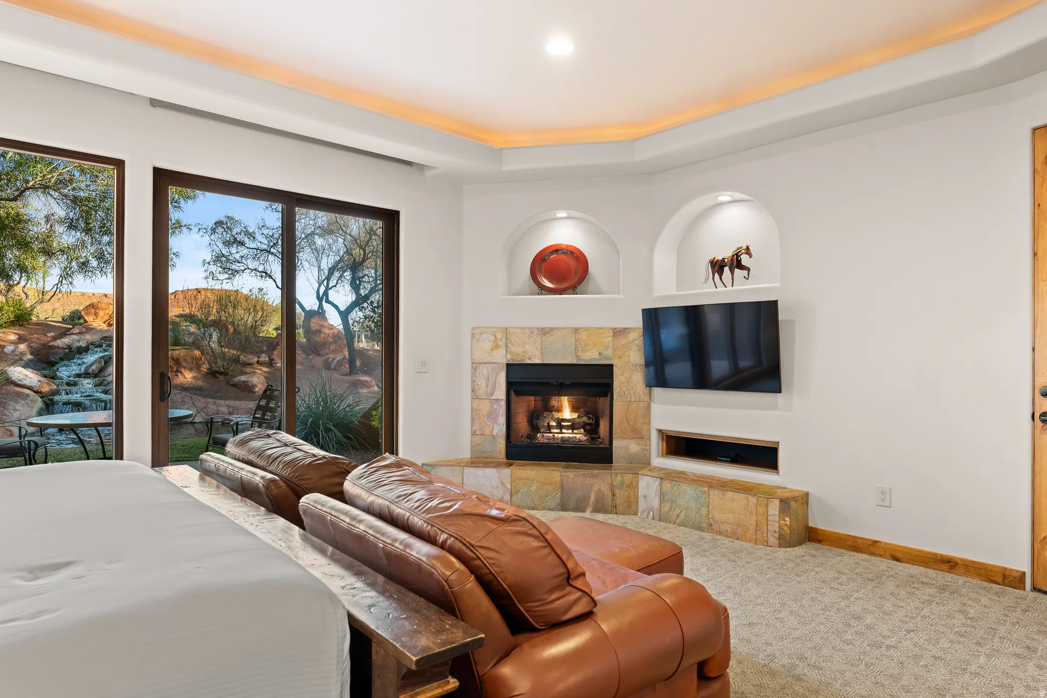 Recessed ceiling lighting, a corner fireplace with stone surround, dual arched wall niches, and sliding glass door access to an outdoor area with a water feature