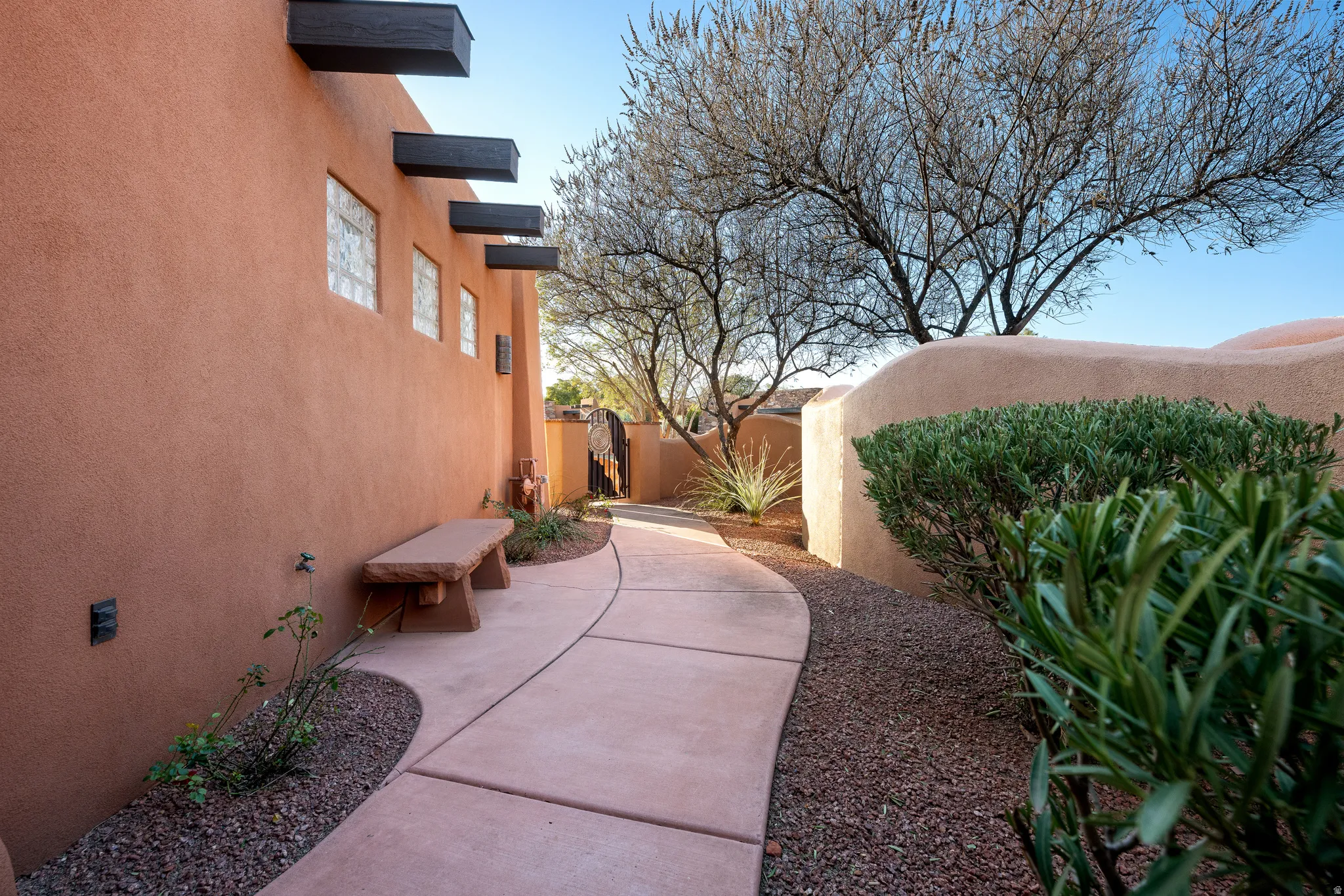Curved pathway with xeriscape landscaping, stucco wall, decorative wooden beams, multiple privacy windows, and a stone bench