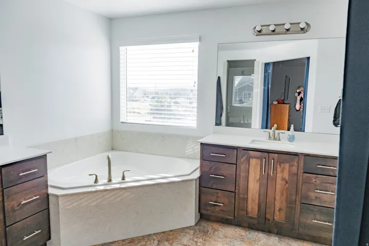 Corner soaking tub with tile surround, dual-tone wood cabinetry vanity with a single basin sink, white countertop, and a large wall-mounted mirror with overhead lighting