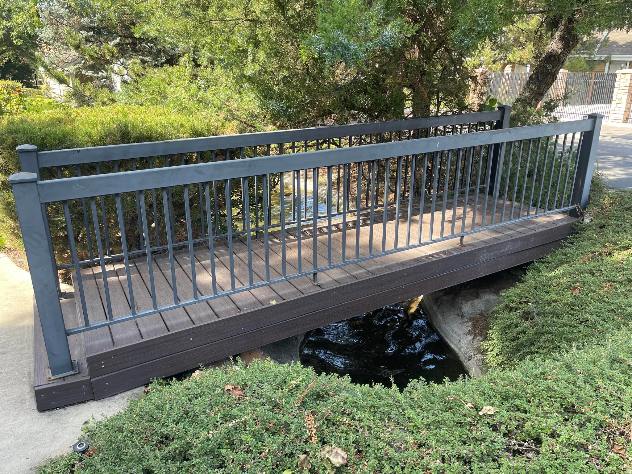 Pedestrian bridge with metal railing and trex decking, surrounded by manicured shrubs