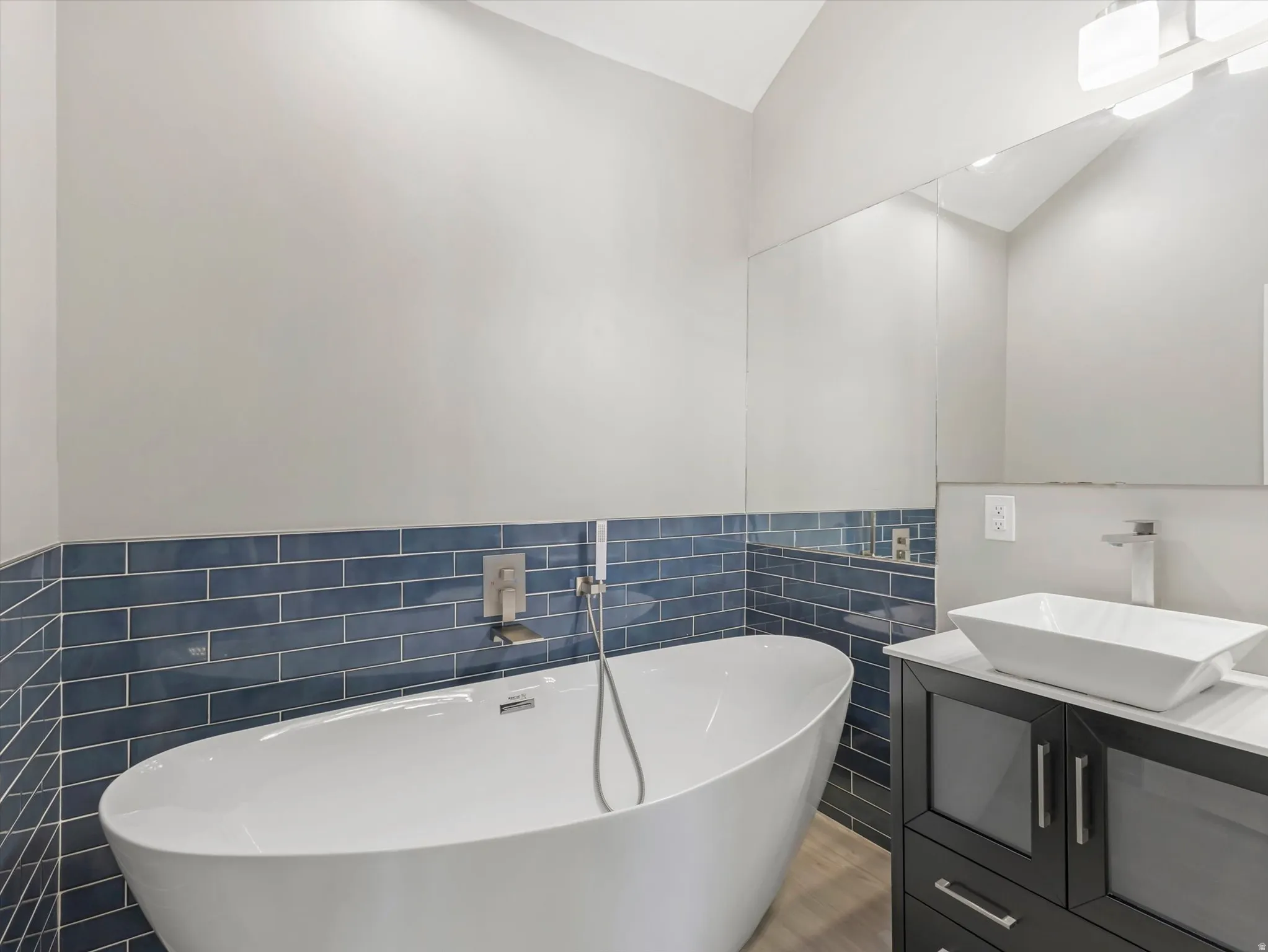 Master Bath with Freestanding soaking tub, blue subway tile accent wall, modern floating vanity with vessel sink, brushed nickel fixtures, Skylight, and wood-finish flooring