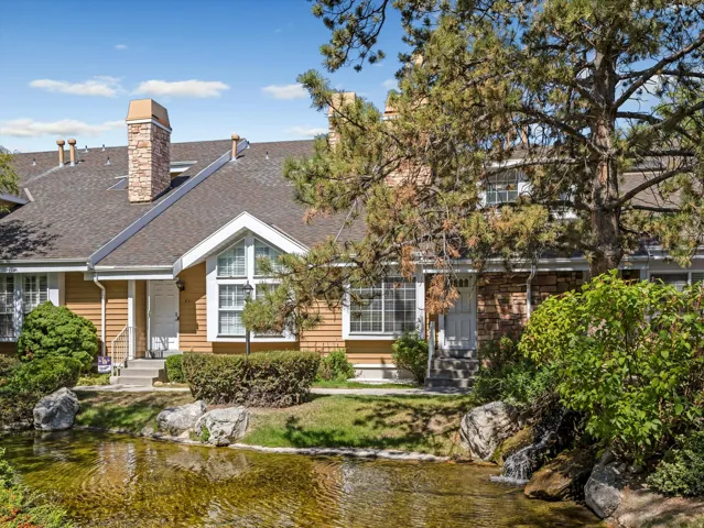 Stone-clad chimney, multiple gabled roofs, decorative peak windows, water feature with rock accents, and lush surrounding landscaping